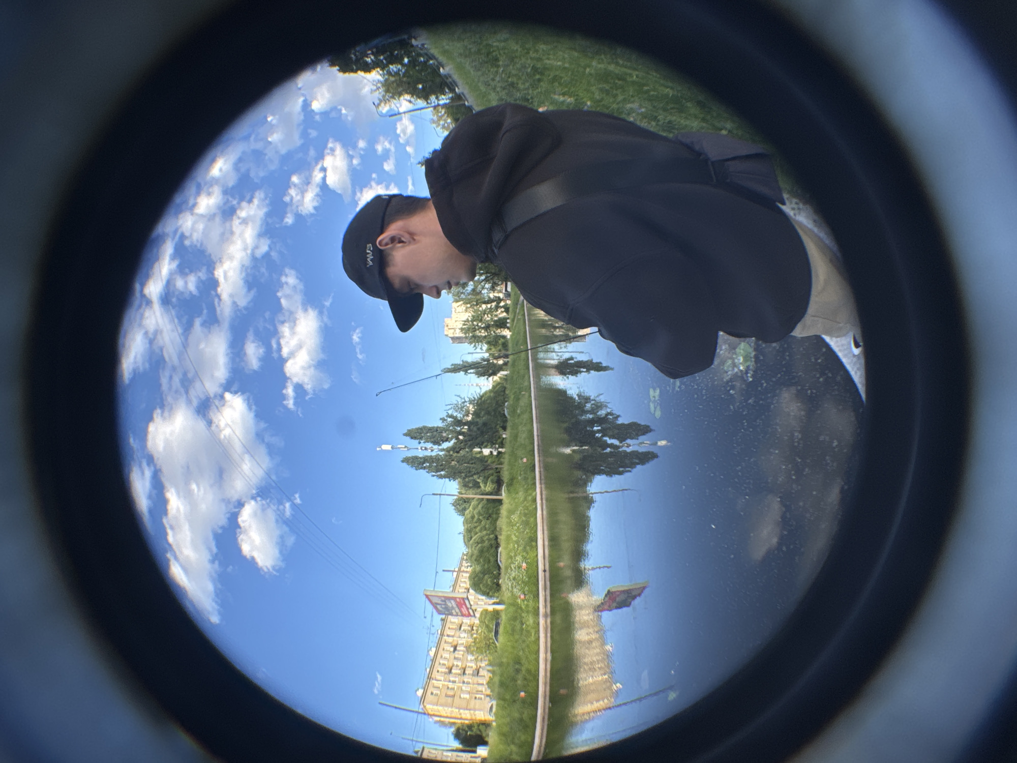 This is a fisheye lens photograph taken in Saint Petersburg, Russia. The image shows a young male wearing a black hoodie, a black baseball cap with a white "VANS" logo, and light-colored pants. He is standing near the edge of a canal or river, appearing to be fishing with a rod. The water reflects the blue sky, white clouds, buildings, and trees on the opposite bank. On the far bank, there are apartment buildings, lush green grass, trees, and streetlights. A billboard with red and white lettering is visible in the background. The foreground is dominated by the dark, circular frame of the fisheye lens, which distorts the scene and creates a sense of looking through a porthole. The overall scene depicts a casual, outdoor activity in an urban environment.