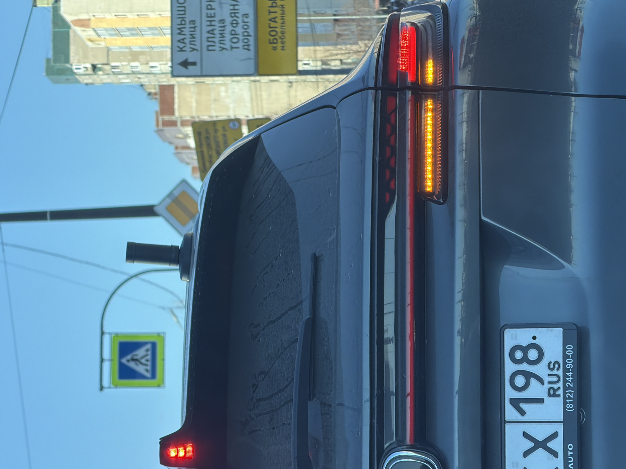 The image depicts the rear view of a dark-colored SUV, likely a crossover or electric vehicle, captured on a clear day in Saint Petersburg, Russia. The vehicle's brake lights are illuminated, displaying a prominent horizontal red LED strip across the tailgate and amber sequential turn signals (though not actively flashing). A cylindrical object, possibly a sensor for advanced driver-assistance systems or a communications antenna, is mounted centrally on the roof. The rear window shows streaks of dried water or dirt, with a single wiper blade resting horizontally.
A Russian license plate is affixed to the lower part of the tailgate, partially obscured but clearly showing "198. RUS," indicating the vehicle is registered in Saint Petersburg and the Leningrad Oblast region. The license plate frame includes a local phone number, (812) 244-90-00, which is also consistent with Saint Petersburg.
In the background, under a bright blue sky, several multi-story residential or commercial buildings are visible. Multiple road signs are present: a square blue sign with a white triangular border and a pedestrian symbol, indicating a pedestrian crossing; a square sign with a yellow diamond on a white background, indicating a main or priority road; and a large white directional sign with an upward arrow pointing to "КАМЫШОВАЯ улица" (Kamyshovaya Street), "ПЛАНЕРНАЯ улица" (Planernaya Street), and "ТОРФЯНАЯ дорога" (Torfyanaya Doroga). Another yellow sign beneath these mentions "«БОГАТЫРЬ» мебельный", referencing a "Bogatyr" furniture store. These street names and the area code confirm the location to be in Saint Petersburg, specifically near the intersection of Kamyshovaya Street and Planernaya Street in the Primorsky District. No individuals or specific actions are discernible beyond the vehicle being stationary or moving slowly in traffic.