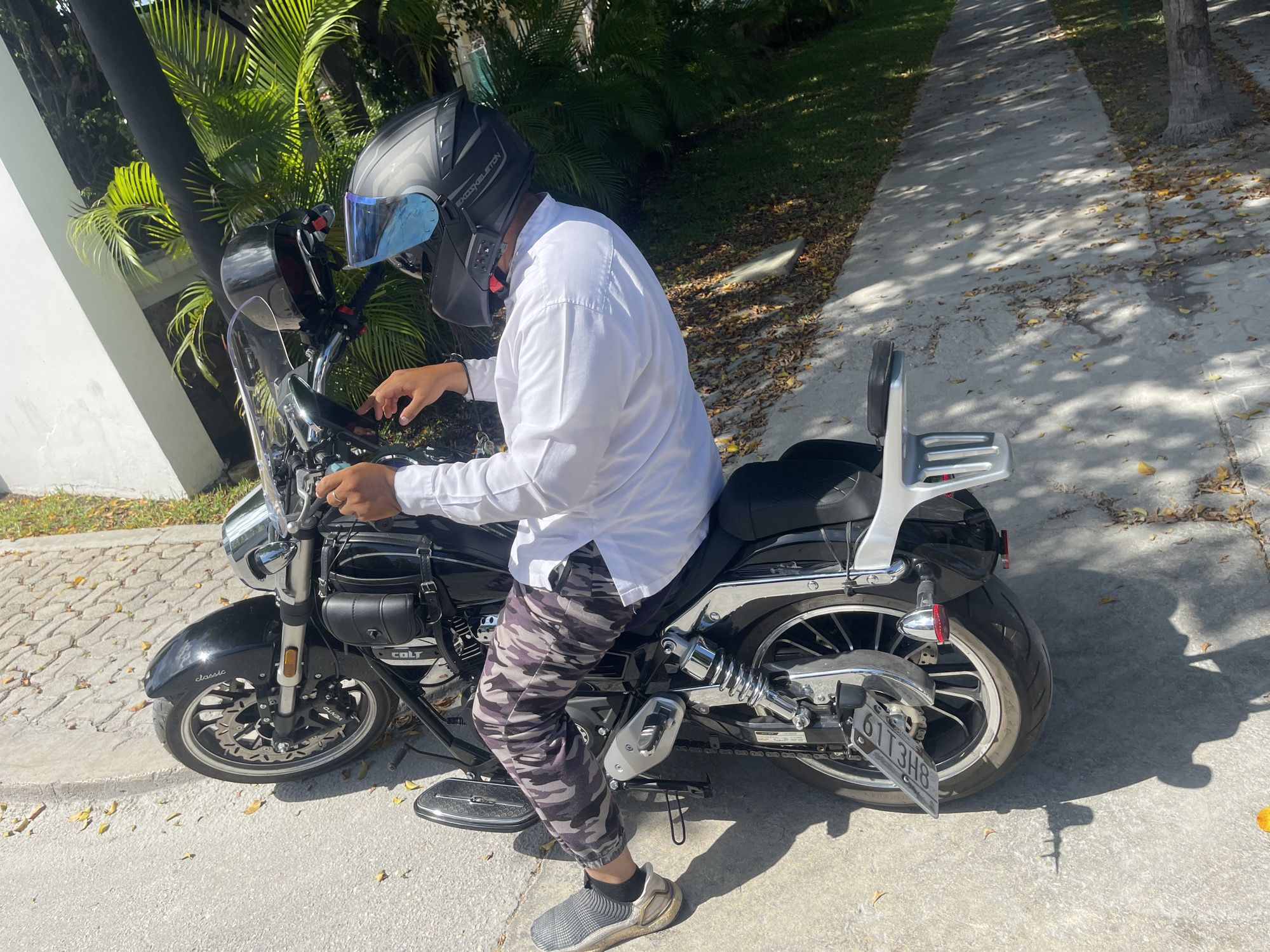 A man wearing a white shirt, camouflage pants, gray sneakers, and a motorcycle helmet is seated on a black Vento Colt motorcycle with a silver luggage rack and a clear windshield. The motorcycle has a license plate that reads "6113H8". The setting appears to be a concrete driveway or sidewalk next to a street in Playa del Carmen, Mexico, with trees and foliage visible in the background. The time of day appears to be sunny.