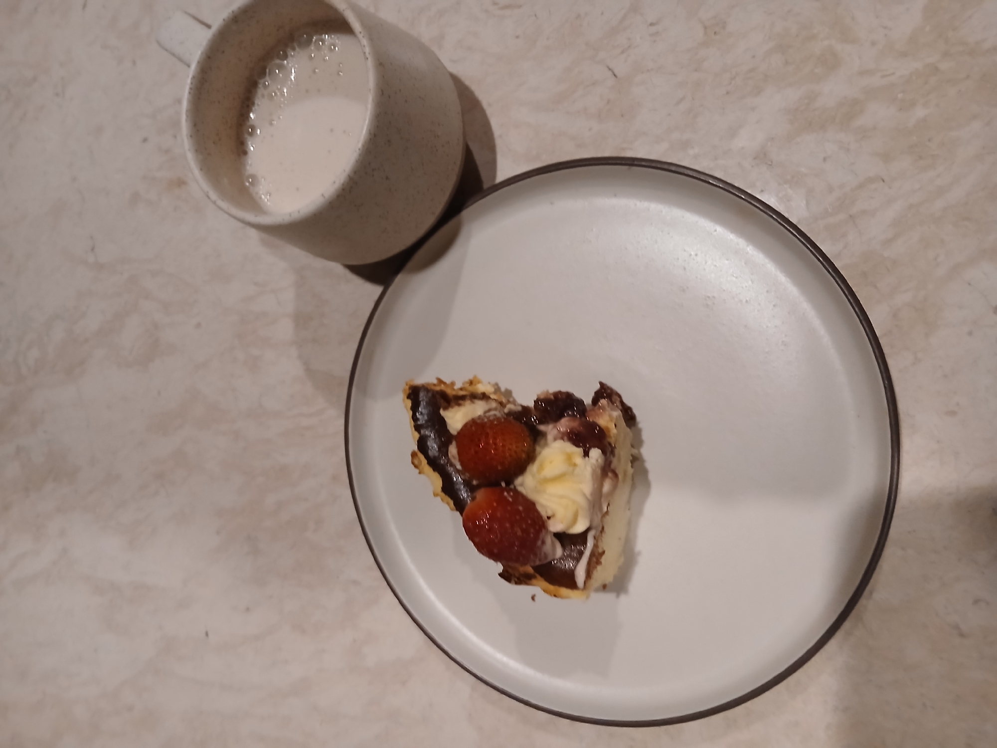 A slice of cheesecake topped with strawberries and cream sits on a white plate with a dark rim. The cheesecake has a dark crust and appears to be a Basque cheesecake due to its caramelized top. Beside the plate, a speckled white mug contains a light-colored liquid, possibly milk or a creamy beverage, with some foam on top. The items are arranged on a countertop with a marbled pattern. The lighting suggests it is either indoors or nighttime, with a shadow cast by the mug. There is no visible text in the image. The scene depicts a dessert and drink, likely a casual snack or meal.