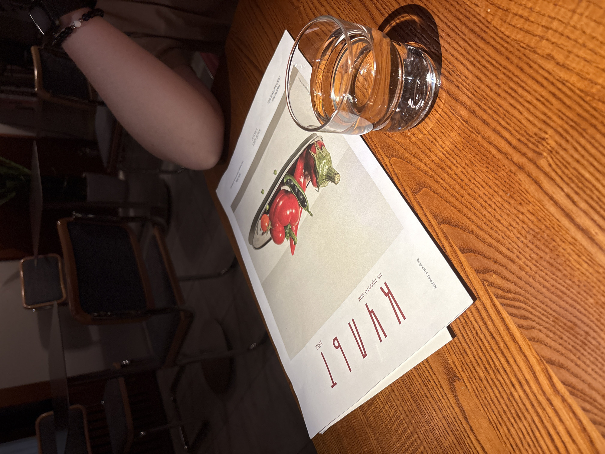The image shows a close-up view of a wooden table in a restaurant. On the table, there is a menu with an illustration of vegetables and a glass of water. In the background, chairs and tables of the restaurant are visible, suggesting a modern, somewhat minimalist interior. The setting appears to be indoors, likely a dining establishment. No specific details or landmarks confirm the location as Rostov-on-Don, Russia.