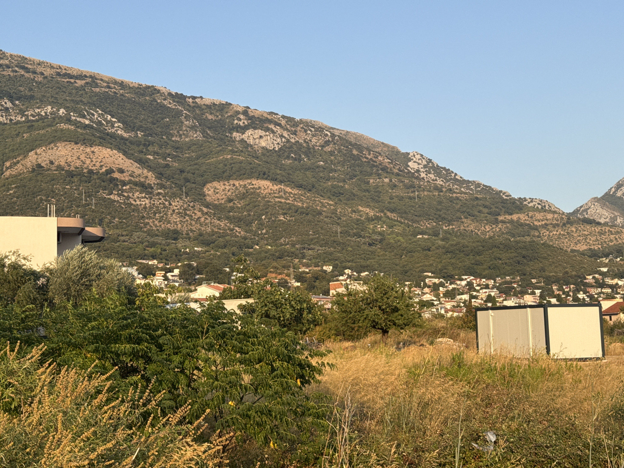 The image depicts a semi-urban landscape with a backdrop of a lush, green mountain range under a clear blue sky. In the foreground, dry, golden grasses and various green shrubs are visible. To the left, the side of a light-colored building with a curved overhang is partially in frame. Further into the scene, a cluster of buildings with red-tiled roofs forms a settlement nestled on the lower slopes of the mountain. Two modular, light-colored structures with dark trim are positioned in the mid-ground amidst the dry grass. The overall scene suggests a warm, sunny day in a mountainous coastal region.