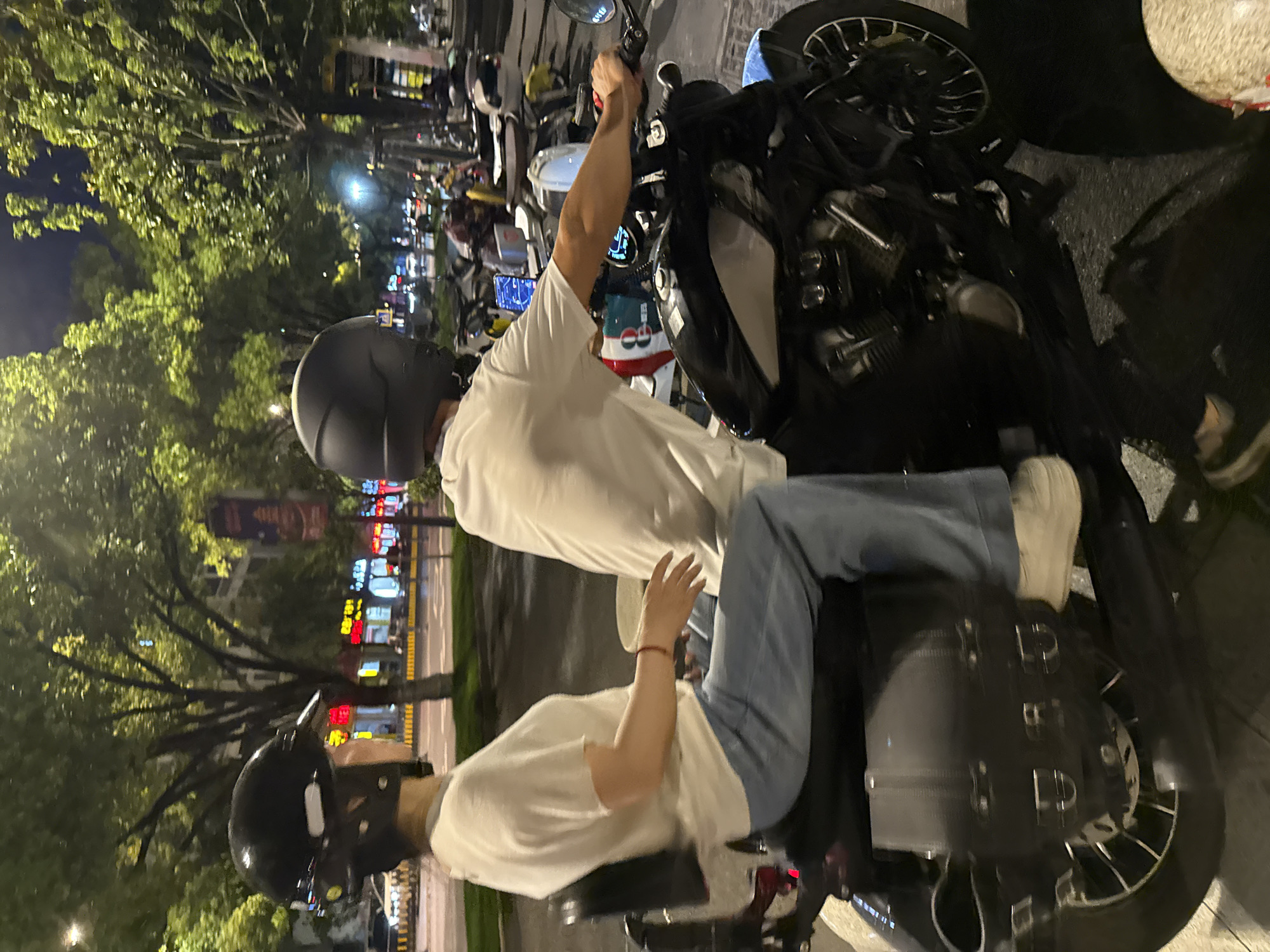 Two individuals, a male and a female, are riding a motorcycle at night in Yiwu, China. The female, wearing a black helmet and a white t-shirt with light blue jeans, is seated behind the male. The male, also wearing a black helmet and a white t-shirt, is operating the motorcycle. The motorcycle is dark-colored with black saddlebags on either side and a blue accent on the rear fender. The background consists of a tree-lined street with illuminated storefronts and other parked motorcycles, suggesting a commercial or pedestrian area in the city. The lighting indicates it is nighttime.