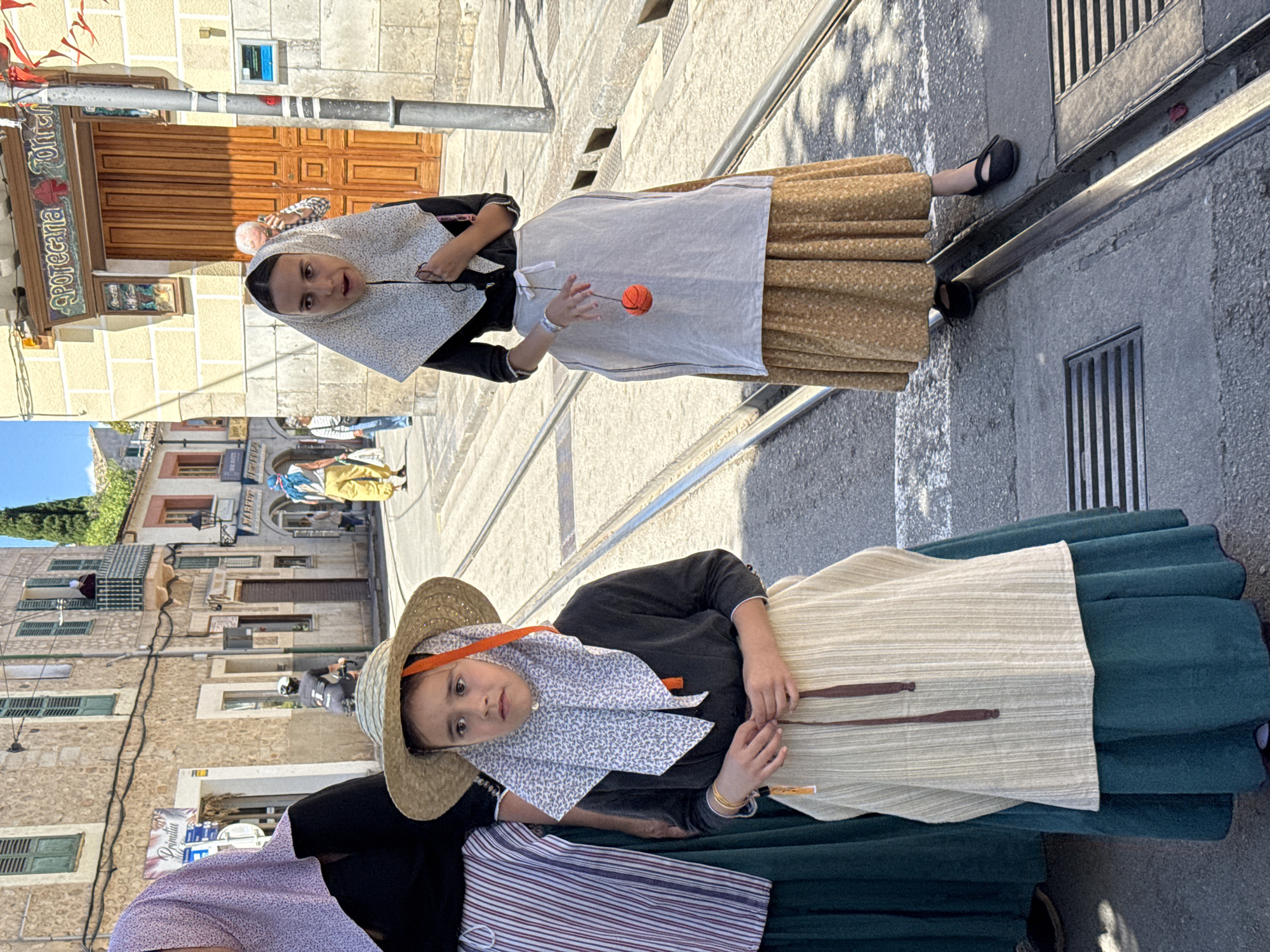 Two young girls in traditional Mallorcan dress stand on a cobblestone street in Sóller, Spain. The first girl wears a brown-patterned skirt, a white apron, and a patterned head covering and plays with a red ball. The second girl wears a green skirt, a white apron, and a straw hat over a patterned head covering. The street has tram rails. The background features buildings typical of Sóller. A pharmacy sign, "Apotecaria Tortell," is visible on one of the buildings. In the distance, more people dressed in similar traditional attire can be seen on the street.