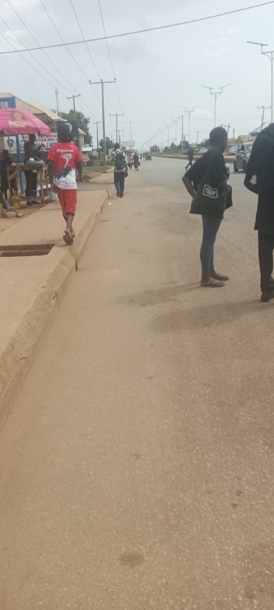The image captures a scene on a street in Bukuru, Nigeria. In the foreground, a person walks on a concrete sidewalk with a drainage channel. This person wears a red shirt with white accents and red shorts. Further down the street, several other individuals are visible, some walking along the road and others appearing to be gathered near a building on the left side of the image. The street itself is paved and has multiple lanes. Power lines and streetlights run along the side of the road, indicating it's a developed urban area. Vehicles are present in the distance, suggesting ongoing traffic. The overall environment appears to be a typical day in a Nigerian city, with a mix of pedestrian and vehicular activity.