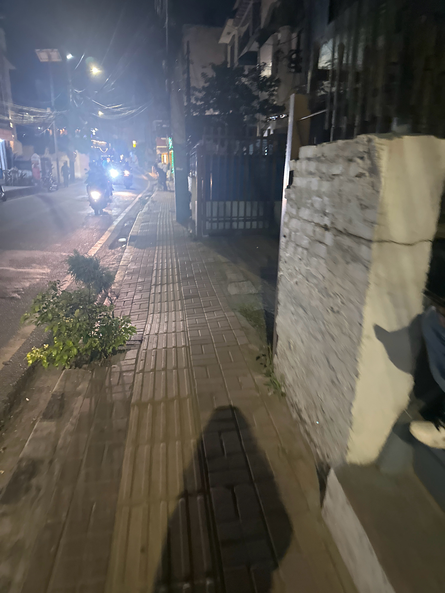 The image captures a nighttime scene on a city street in Lalitpur, Nepal. A paved sidewalk runs along the right side of the frame, with textured tactile paving tiles indicating a pedestrian walkway. To the left, a street is visible, illuminated by streetlights and the headlights of a motorcycle, which is the prominent subject in the foreground of the street. Several other motorcycles are also present on the road, some with riders. Buildings line the street, and wires are strung overhead. On the right side of the sidewalk, a section of a rough, unfinished brick wall is prominent, with a person's legs and feet visible at the bottom right, seated on a step. A shadow of the photographer is cast on the sidewalk. The environment appears wet, possibly from recent rain, with reflections visible on the pavement. A small bush grows in a concrete planter by the side of the road.