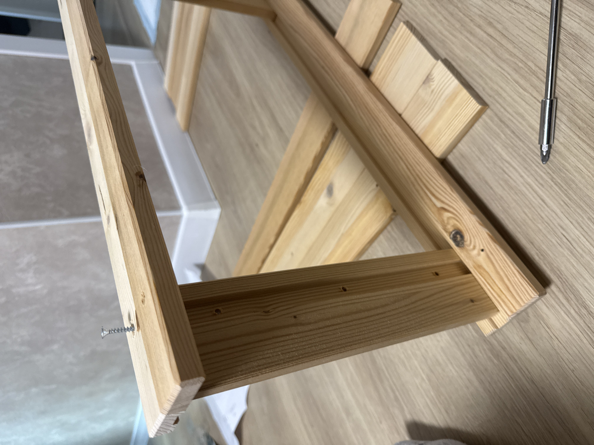 The image depicts a partially assembled wooden structure on a light-colored wooden floor, possibly indoors. The structure is constructed from light-colored wood and appears to be held together with screws. A screwdriver is lying on the floor nearby. The location is indeterminate and unspecified.