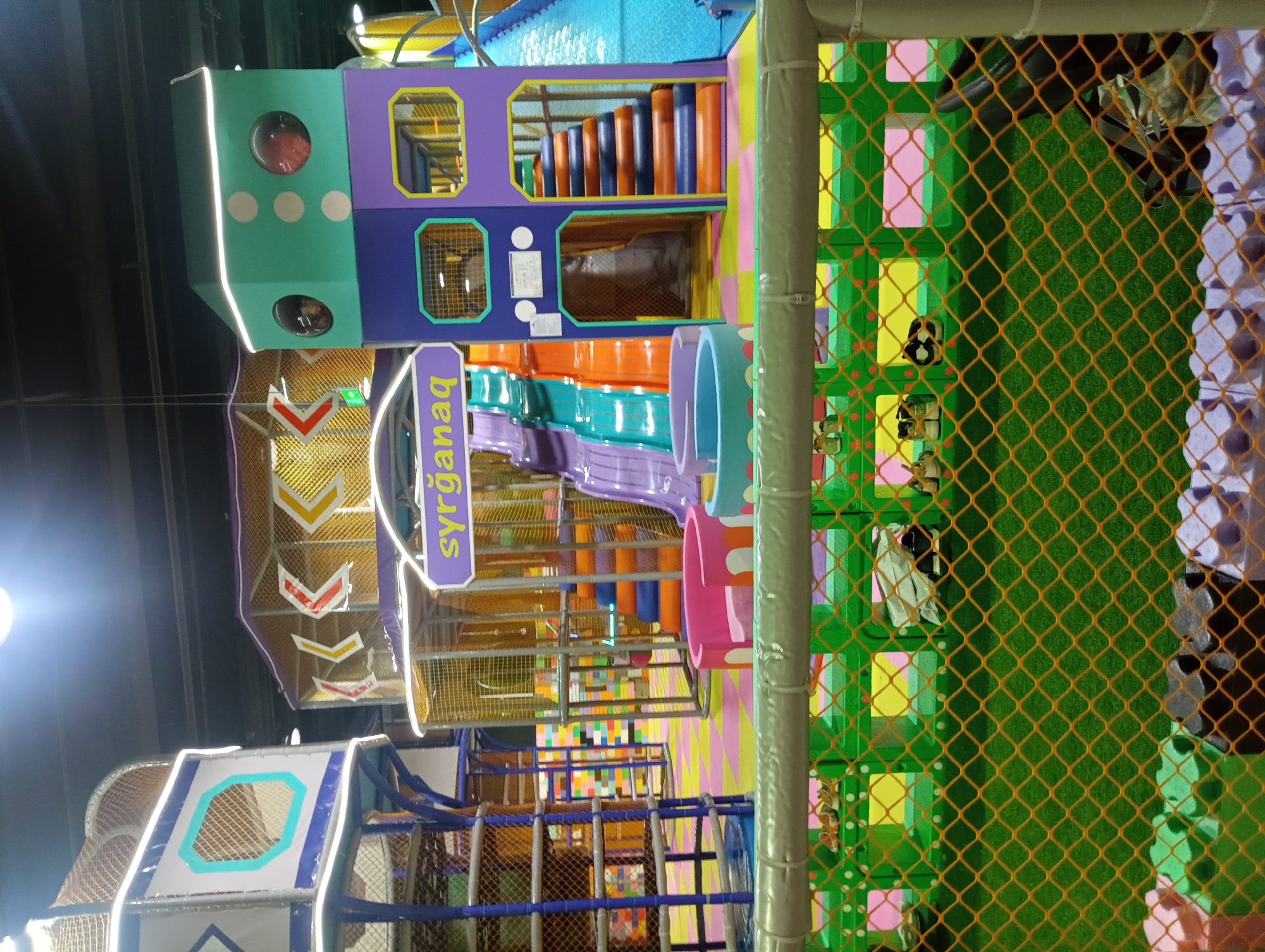 The image depicts an indoor children's play area, likely a playground or soft play center, at an unspecified location in Almaty, Kazakhstan. The environment is brightly colored with various play structures. In the center, there are large, multi-level play structures with slides, climbing nets, and staircases. One structure is predominantly purple and green, featuring a sign with the text "Syrganaq," which translates to "carousel" or "merry-go-round" in Kazakh, though the structure itself does not appear to be a carousel. Other play elements include a purple curved seating area and a blue cylindrical soft play structure. The floor is a patchwork of colored padded mats in yellow, pink, and green. In the foreground, a green safety fence with an orange net encloses a grassy-looking surface, and on a shelf behind the fence, several pairs of children's shoes are visible. The lighting suggests artificial indoor illumination. There are no visible people engaged in play, but the presence of shoes indicates that children have been or are present in the facility. The overall impression is one of a safe and stimulating environment designed for children's entertainment.