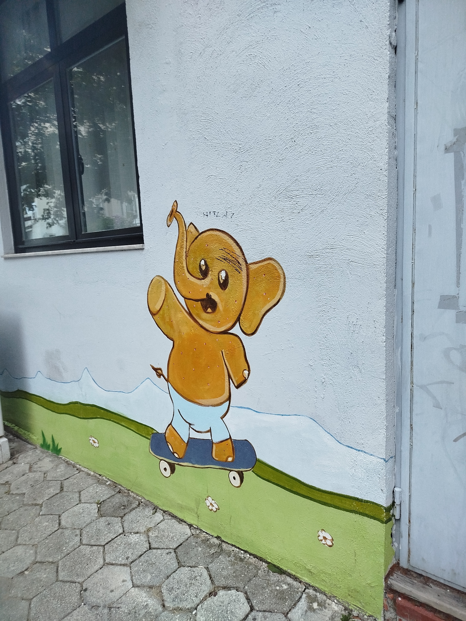 The image shows a mural of a cartoon elephant on a white textured wall. The elephant is yellow with a blue diaper, riding a skateboard. It has its trunk raised and one arm outstretched as if waving. The mural also depicts a green grassy area with a few small white flowers and a wavy blue line representing mountains in the background. A dark-framed window is visible to the left of the mural. The wall is next to a light-colored doorframe and a concrete walkway made of hexagonal pavers. The overall scene is outdoors, suggesting it is part of a building's exterior, likely in a public space. The location in Omiš, Croatia, is identified as being on the exterior wall of a building near a school or kindergarten, given the childlike nature of the artwork and the presence of a window that could be for a classroom.