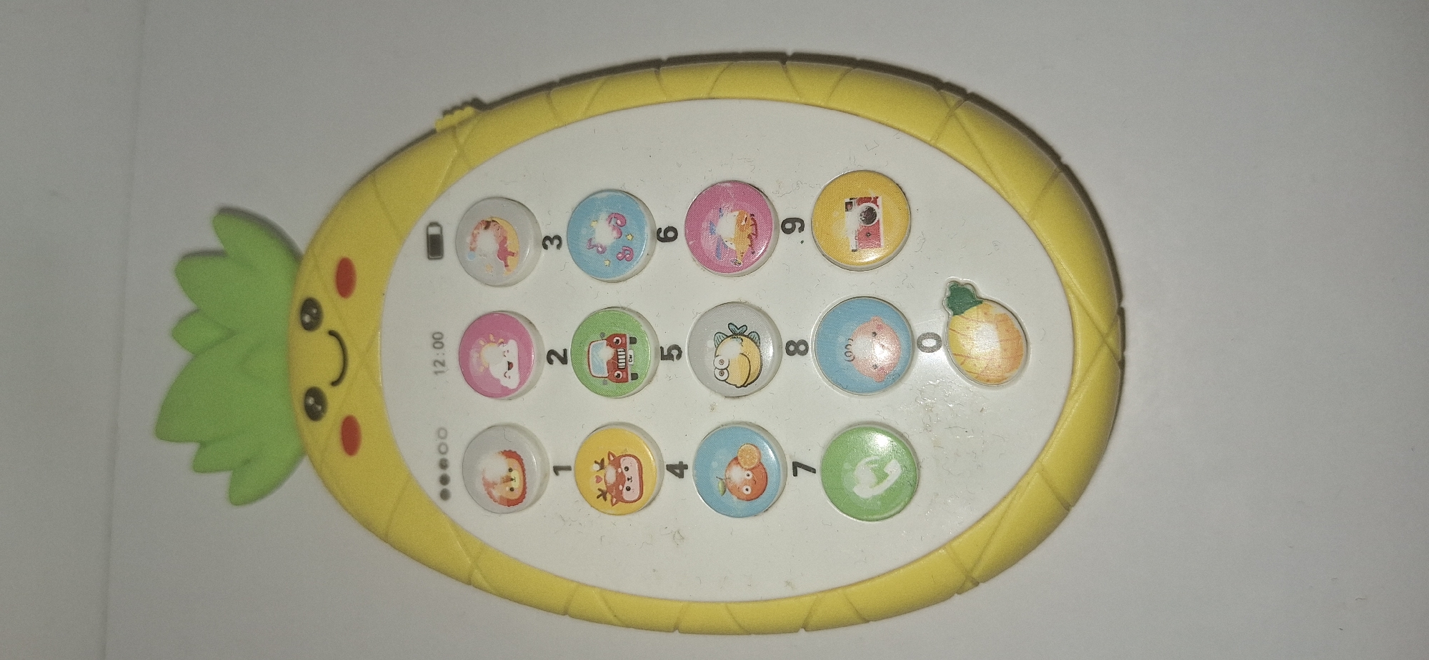 The image shows a toy pineapple-shaped phone. The phone is yellow with a green leafy crown and a smiling face. It has a grid of circular buttons with various cartoon images and numbers. The buttons depict animals, vehicles, celestial bodies, and a camera. The time displayed on the phone's screen is 12:00. The phone is lying on a white surface. There are no discernible details indicating its location in Bila Tserkva, Ukraine.