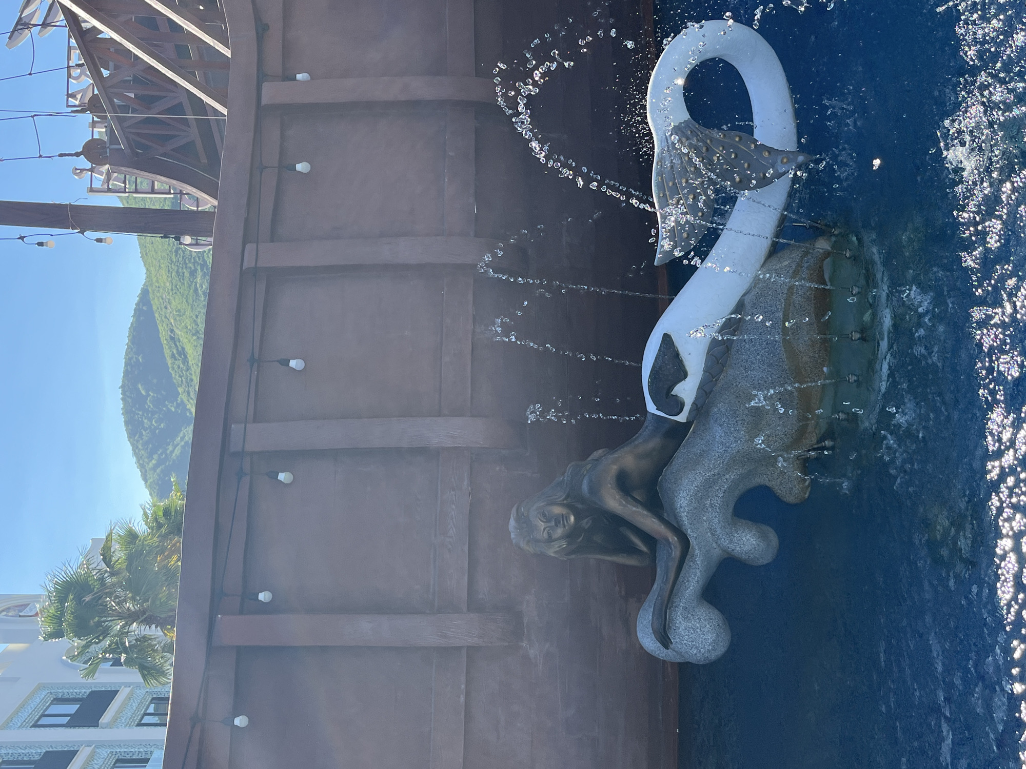 This media file displays a fountain sculpture with a swan and a mermaid, set against a backdrop of a wooden wall and a lush, green mountain range. The swan sculpture is white with grey feathered details on its wings, and water sprays from its neck and tail, creating a shimmering effect. The mermaid, cast in bronze, is positioned at the base of the swan, her head tilted back as if enjoying the water.
The scene is illuminated by bright sunlight, suggesting it is daytime, likely mid-morning or afternoon given the clear blue sky and the shadows cast. The presence of palm trees and modern white buildings in the background hints at a tropical or sub-tropical coastal location. The overall atmosphere is one of tranquility and artistic beauty, with the water from the fountain adding a dynamic and refreshing element to the scene. The specific location is Bac Nha Trang Ward, Vietnam. There is no visible text in the image.