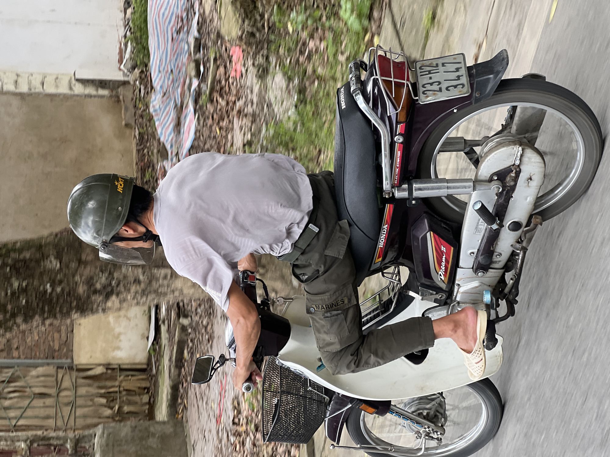 In a medium shot, a person rides a purple Honda Dream II motorcycle on a paved road in Hà Giang 2 Ward, Vietnam. The rider, viewed from the rear-right, wears a green helmet with a clear face shield and a light gray t-shirt. Their dark green cargo pants are adorned with a "MARINES" patch. They are also wearing light-colored slip-on sandals. The motorcycle has a black basket on the front and a luggage rack at the back. A license plate reading "23-H2 3447" is visible. The background shows a tree with rough bark, a weathered wall, and some foliage, with a striped tarp partially visible to the right. The scene suggests a typical street scene in Vietnam.