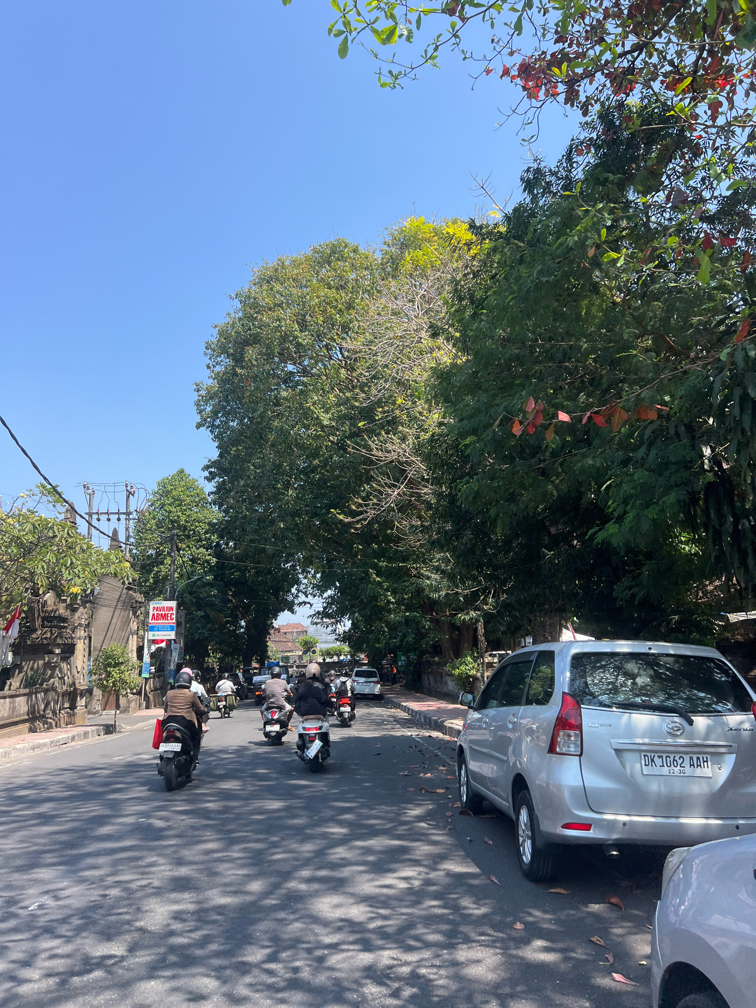 The image depicts a sun-drenched street scene in Denpasar, Indonesia, on a clear day. A silver minivan, identified by the license plate DK 1062 AAH, is parked on the right side of the road, partially obscuring another vehicle. The road is lined with lush green trees casting dappled shadows on the asphalt. Several people are seen riding motorbikes, navigating the street. On the left side of the image, a traditional Balinese stone wall runs alongside the road, with a red and white Indonesian flag visible. A sign for "PAVILIUN ABMEC" is also present. Electrical poles and wires are visible in the background, suggesting urban infrastructure. The overall atmosphere is one of a typical day in a tropical city.