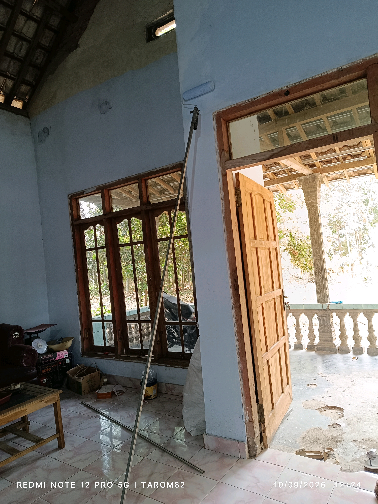 The image depicts an interior scene of a house, likely undergoing renovation or cleaning, in Rejoagung, Indonesia. The room has light blue walls and a tiled floor. In the foreground, a wooden table with a glass inlay is visible, along with various items like a scale, a generator, and boxes. A long metal pole, possibly for painting or cleaning, extends from the floor upwards towards the ceiling, where a paint roller is attached. A large wooden door is ajar, leading to an outdoor area with a porch supported by decorative pillars and a balustrade. Outside, trees and sunlight suggest a daytime setting. The image is dated 10/09/2026.