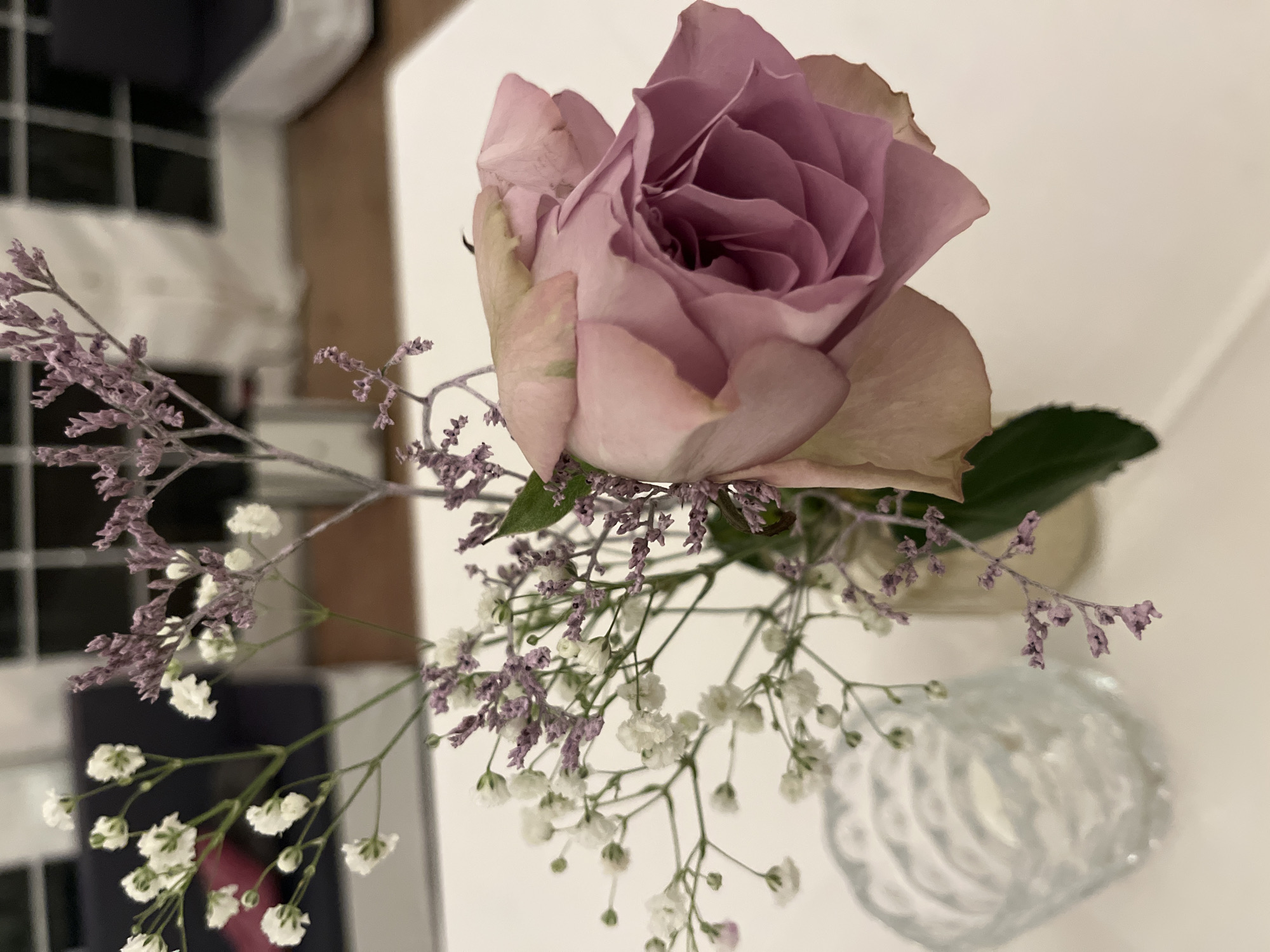 A top-down view shows a single light purple rose in a small, clear glass vase, accompanied by small purple and white flowers. The arrangement is likely on a white surface. In the background, out of focus, are black and white tiled walls and what appears to be a white wall or surface. The specific location is not discernible from the image. Therefore, it is not possible to confirm this photo was taken in Saarbrücken, Germany.