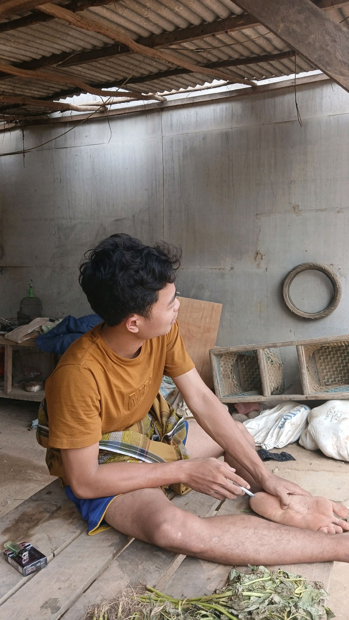 A young man with dark, curly hair is sitting on a wooden platform, facing to his right. He is wearing a mustard-yellow t-shirt and a patterned sarong over blue shorts. His right arm is extended, and he is holding a cigarette between his fingers, appearing to light it with a lighter. His left hand rests on his left knee. Behind him, the scene is a dimly lit, somewhat dilapidated structure with a corrugated metal roof supported by wooden beams. The wall behind him is a rough concrete surface. To his left, there is a wooden table with various items, including a birdcage. To his right, there are woven bamboo shelves and several white sacks. In the foreground, at the man's feet, is a pack of cigarettes and some green leafy plants. The overall impression is a candid snapshot of a moment of relaxation in a rural or semi-rural setting, possibly in Adi Luhur, Indonesia, given the context.