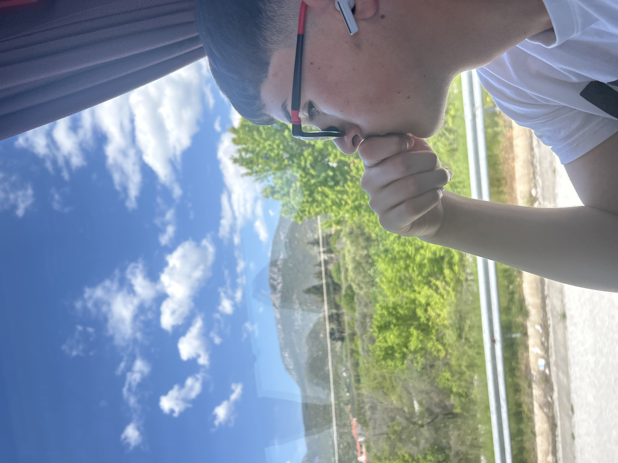 A man with black hair and glasses, wearing a white t-shirt, is looking out of a window. He's holding his fist to his mouth. Outside the window is a view of Gravouna, Greece, featuring a blue sky with clouds, a mountain, and green trees. A highway is also visible through the window. A gray wireless earbud is in the man's ear. Part of a curtain is also visible at the top of the image.