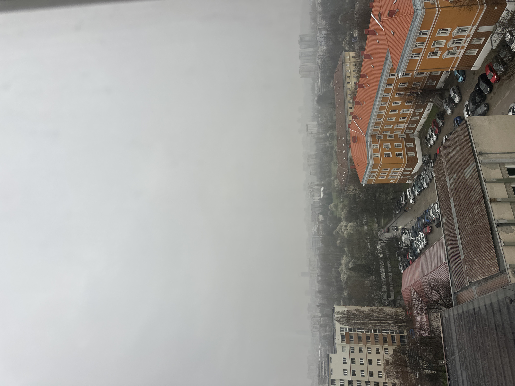The image presents a high-angle view of Sofia, Bulgaria, captured on a foggy day. The sky is overcast, obscuring the background and distant buildings. The foreground consists of residential buildings with orange tiled roofs and several parked cars in the adjacent parking lots. The cityscape gradually fades into the fog, with only faint outlines of buildings visible in the distance. There are no people visible. The general impression is a hazy, obscured urban landscape.