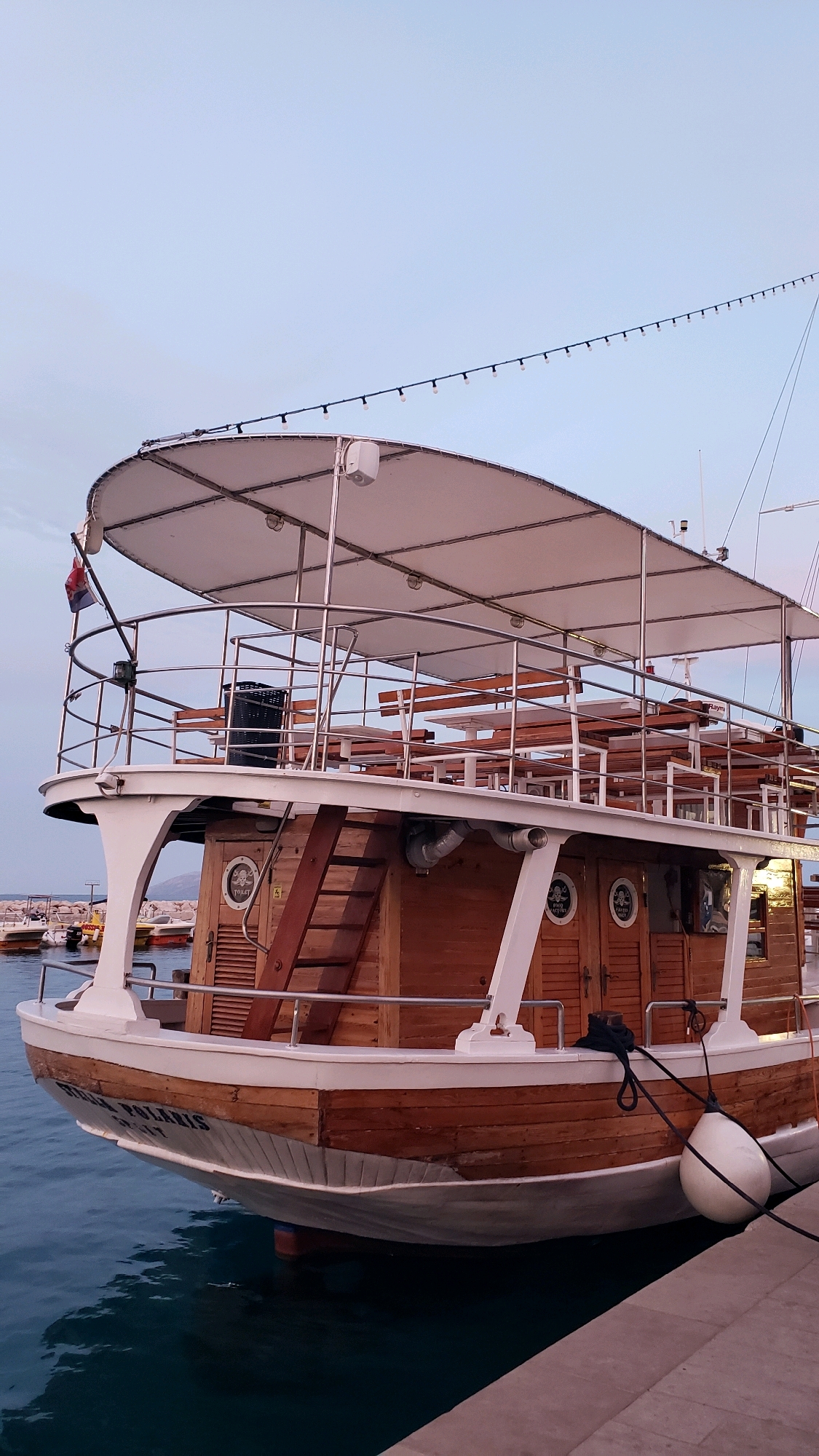 A wooden boat with multiple decks is docked in Općina Tučepi, Croatia. The boat has a white canopy supported by metal poles covering the upper deck, which is furnished with wooden benches and tables. A metal railing encircles the upper deck. A wooden staircase leads to a lower level of the boat, which features wooden doors with circular windows. The boat's hull is painted white and features the name "TEMA POLARIS" in black lettering. A white fender is attached to the side of the boat, and it is secured by a black rope to a pier. The water surrounding the boat is a deep blue, and the sky above is a pale blue with a hint of pink. In the background, other boats are visible docked in the harbor, along with buildings on the shore. The scene appears to be set during the late afternoon or early evening.