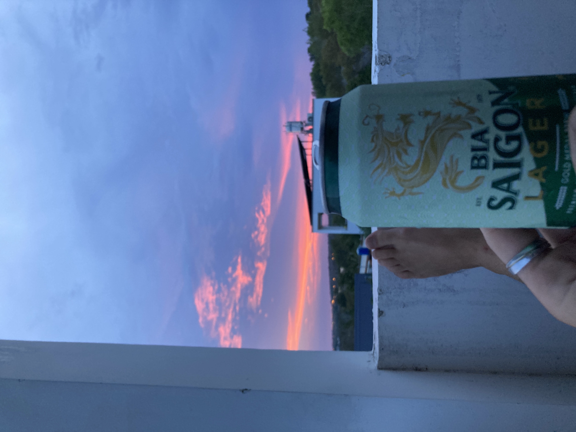 The image shows a can of Bia Saigon Lager held up against a twilight sky. The sky is a mix of blue and pink with scattered clouds. In the background, there is a rooftop structure with a white railing. Trees and possibly buildings are visible in the distance. The location could be Mũi Né, Vietnam, given the beer brand, but there are no unique architectural or geographical features definitively confirming this. A person's arm and foot are partially visible, holding the can.
