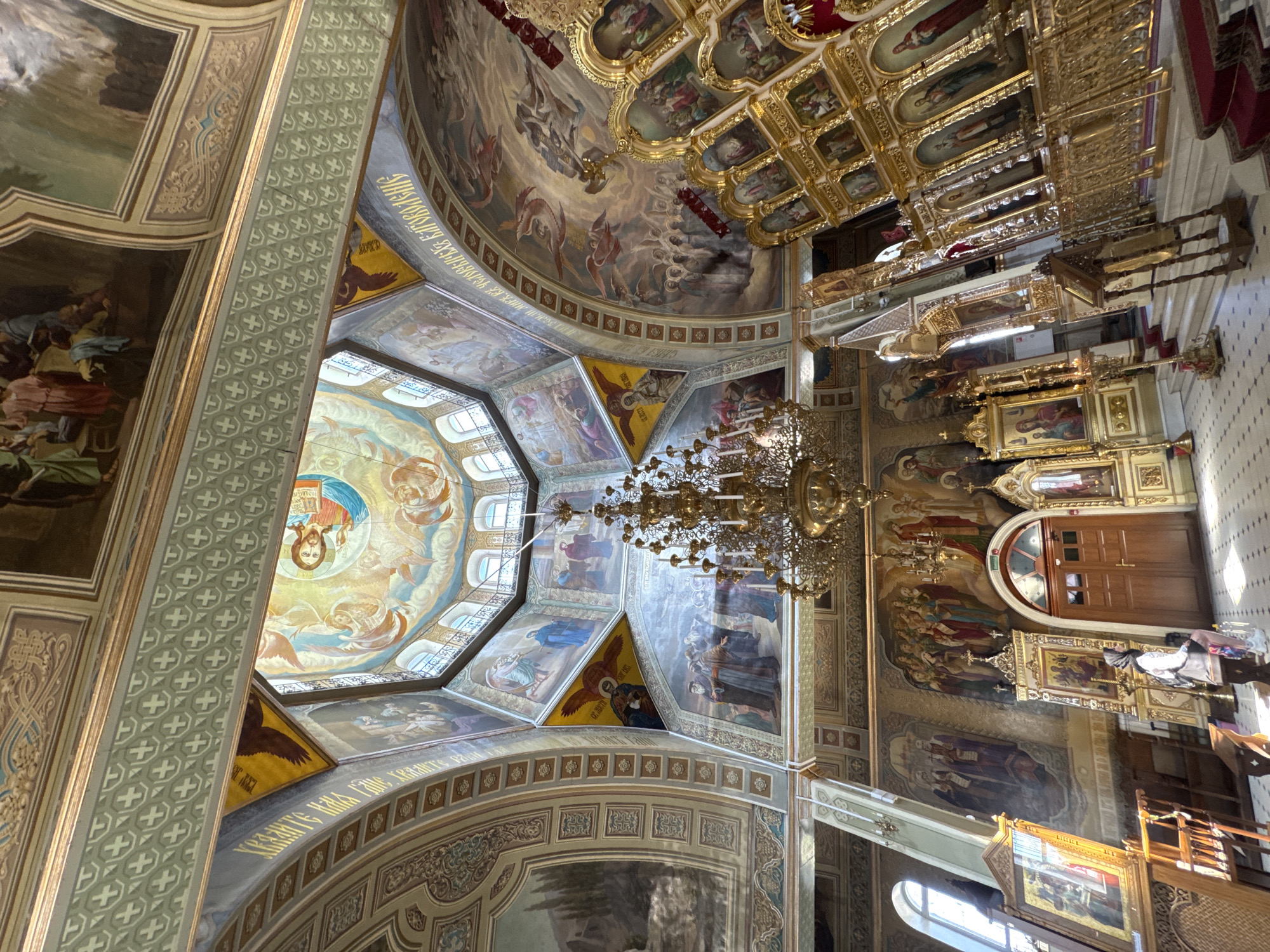 An interior view of an ornate church, likely Russian Orthodox, showcases its rich frescoes and gilded iconostasis. The vaulted ceiling is adorned with depictions of religious figures and biblical scenes, including a prominent fresco of Jesus Christ in the central dome, surrounded by angels. A large, multi-tiered chandelier hangs from the ceiling, illuminating the space. The iconostasis, a highly decorated screen separating the nave from the sanctuary, is made of gold and features numerous framed icons. Several people are present in the church, some of whom appear to be tourists observing the architecture and artwork. The floor is tiled with a mosaic pattern.