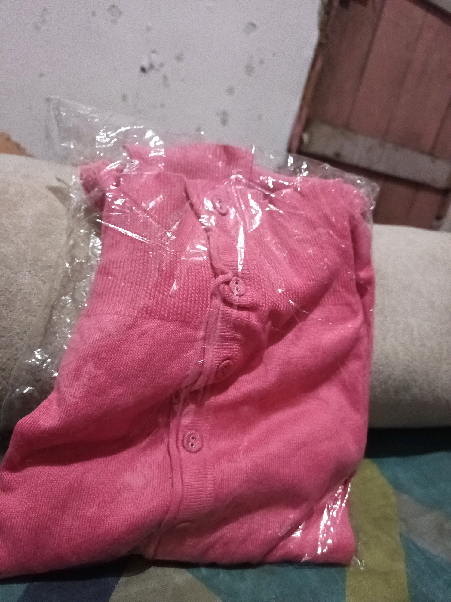 A folded pink cardigan is encased in a clear plastic bag. The cardigan has several buttons down the front. The bag is placed on a light-colored surface, possibly a couch or bed, with a textured, dark-colored surface underneath. In the background, a wall with peeling paint is visible, along with what appears to be a wooden structure or furniture. The scene suggests an indoor setting, likely a home, in South Jakarta, Indonesia.