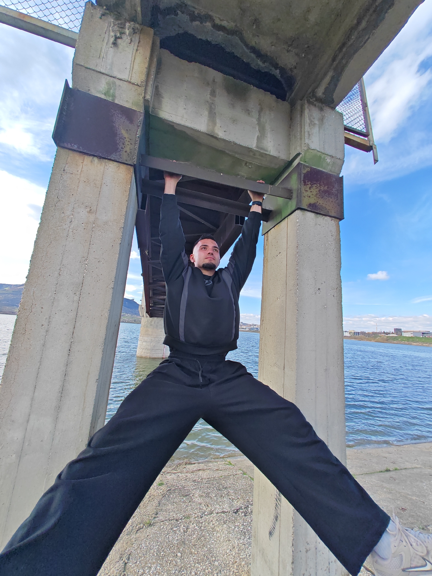A man is performing a gymnastic-style exercise under a bridge. He is holding onto a metal beam with both hands, his arms fully extended, and his legs are spread wide apart, with one foot resting on the base of a concrete pillar. The man is wearing a black sweatshirt, black pants, and sneakers. The bridge is made of concrete and metal, and the surrounding environment includes a body of water, possibly a river or lake, and a clear, blue sky with some clouds. The location is Shumen, Bulgaria, as indicated by the presence of a bridge over water, which is common in the area.