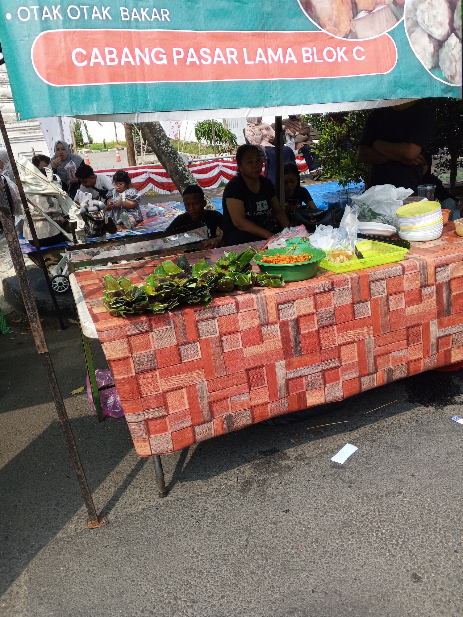 In Serang, Indonesia, a street food stall displays various prepared foods. A red and white patterned tablecloth covers the table, on which are stacked green banana leaf-wrapped parcels, a green bowl filled with what appears to be satay, and a yellow plastic container with other food items. In the background, several people, including women and children, are seated on the ground. A banner above the stall reads "OTAK OTAK BAKAR CABANG PASAR LAMA BLOK C," indicating the type of food sold and its branch location. The scene appears to be outdoors, possibly on a street or market, with a concrete ground and visible shadows.