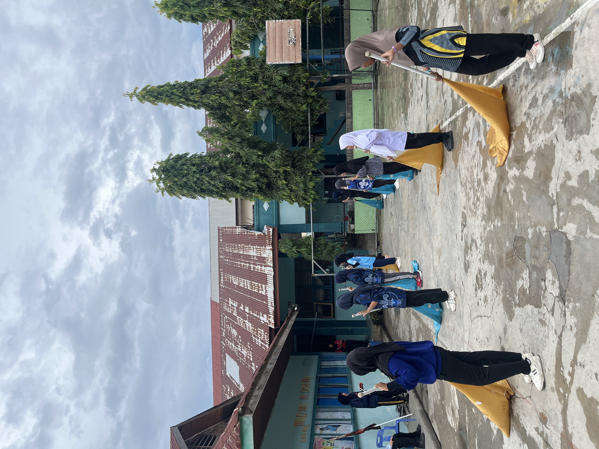 The image depicts an outdoor scene on a cloudy day, likely at a school in Lhokseumawe, Indonesia, given the presence of what appears to be a school building with Indonesian signage visible. A group of young people, predominantly female and wearing hijabs, are arranged in a line on a wet, paved courtyard. They are holding flagpoles with fabric attached, resembling a color guard or performance group. Some are in athletic attire, while others are in more formal school uniforms. In the background, there are several tall, green trees and the facade of a building with a weathered, red corrugated metal roof. A basketball hoop is also visible. The overall atmosphere suggests a rehearsal or practice session.