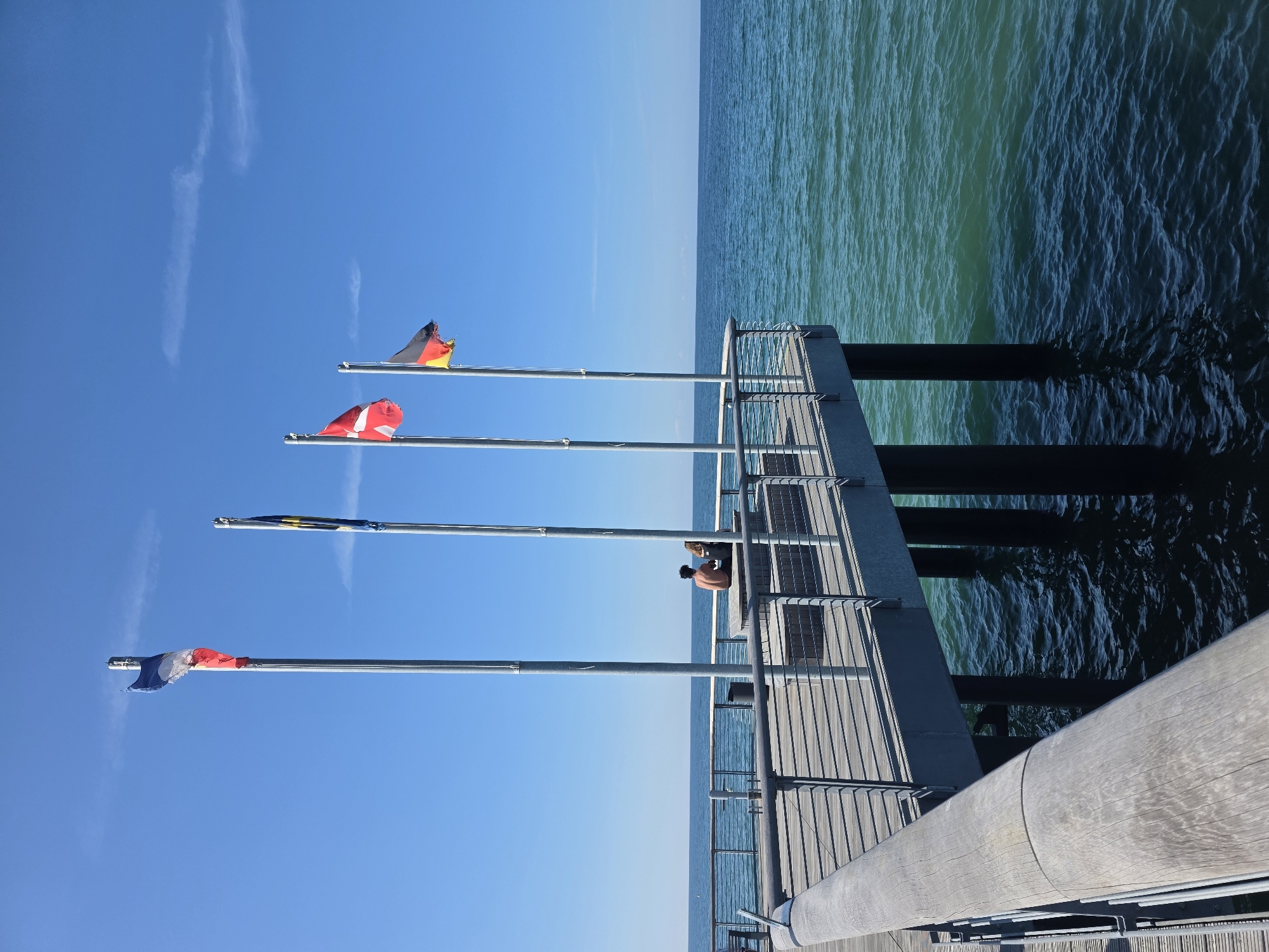 This image depicts a pier extending into the ocean. Four flags fly from poles on the pier, with a clear blue sky above. The flags appear to be the French, European Union, Danish, and German flags. On the pier, two individuals, one with bare shoulders, are seated on a bench, facing the ocean. The wooden pier has metal railings. The ocean water is a deep blue and green, with slight ripples. The scene is set in the coastal region of Wangels, Germany, specifically at the pier in the municipality of Wallnau, which is part of Wangels.
