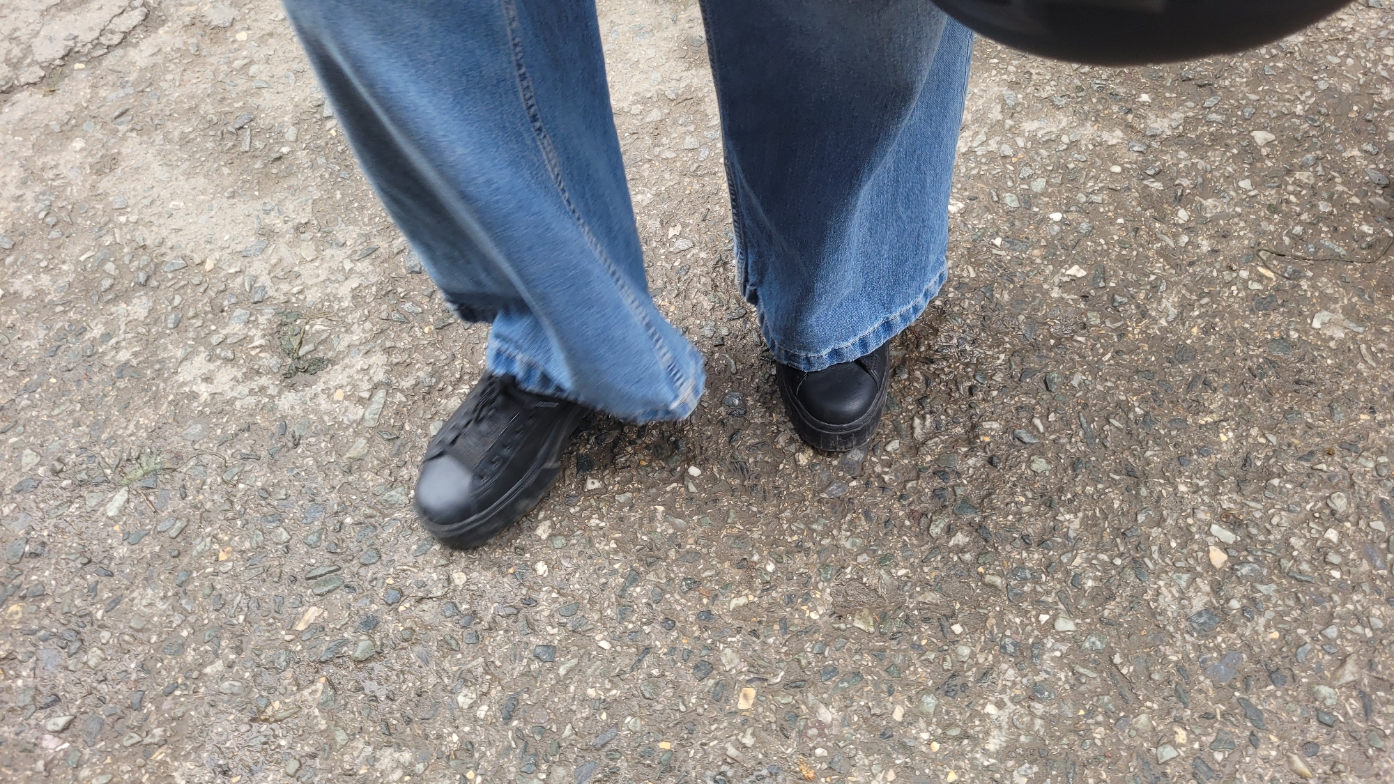 An overhead shot shows a person's legs and feet wearing blue denim jeans and black shoes. The shoes appear to be a lace-up style with a thick sole. The person is standing on a wet, gray asphalt surface that is speckled with small stones and pebbles. The overall scene suggests an outdoor urban environment.