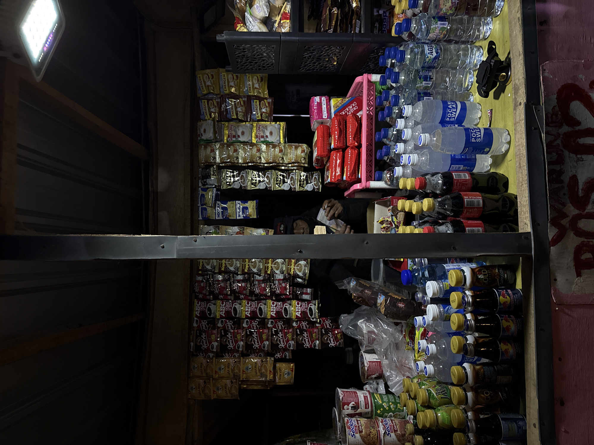 A well-lit, small convenience store in an unidentified location in Indonesia is stacked with various consumer goods on shelves. The shelves are filled with various instant coffee brands such as 'Kapal Api' and 'ABC Plus', as well as a row of 'TOP' flavored instant drinks, bottled water from brands like 'AQUA' and 'POCARI SWEAT', and energy drinks in bottles. The store is possibly managed by a person visible through the shelves, engaged in handling what appears to be cash or documents. The exterior wall of the store has some form of pinkish graffiti.