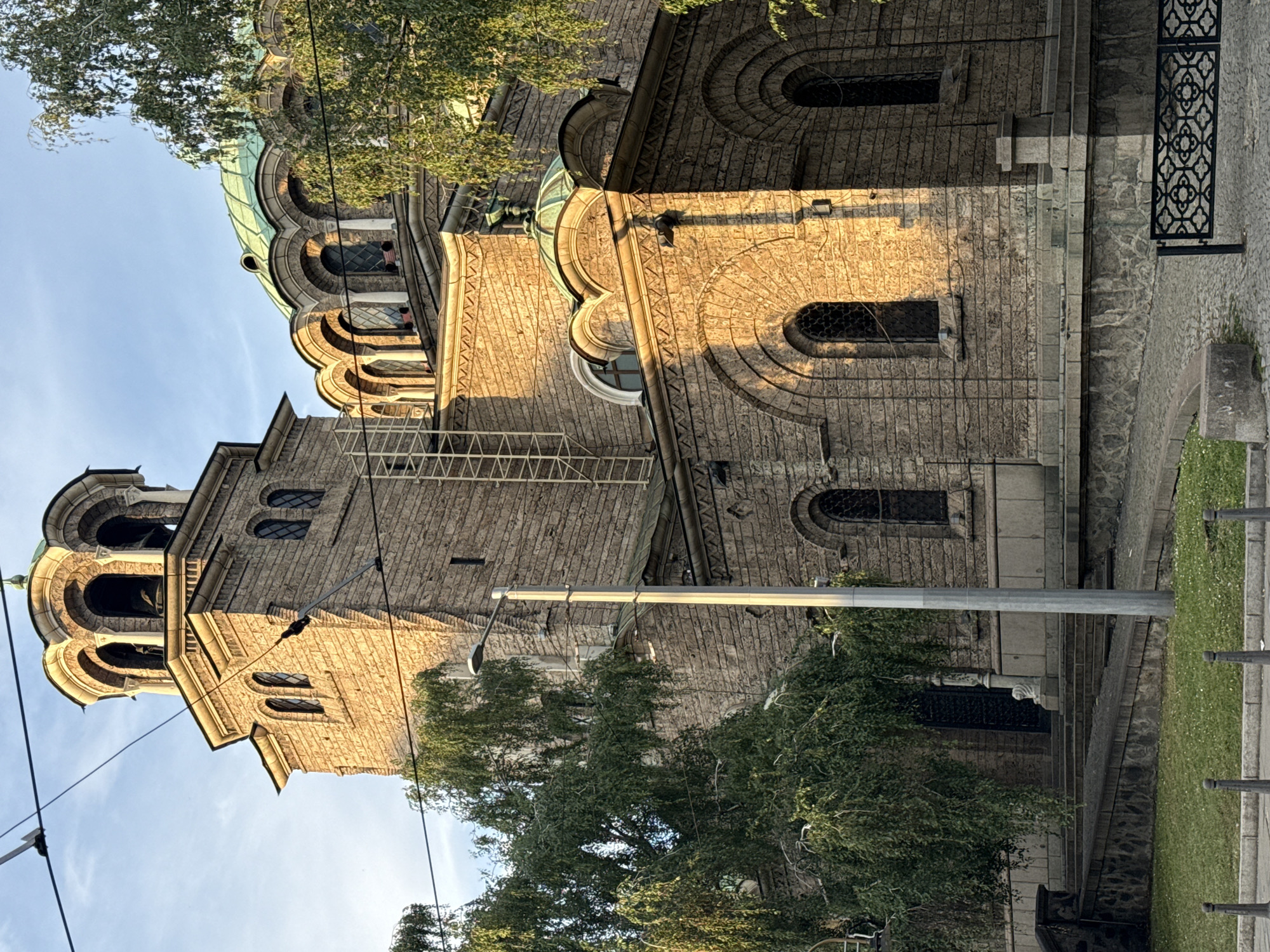 This is a photograph of the Alexander Nevsky Cathedral in Sofia, Bulgaria, taken from a low angle, with the sun illuminating the right side of the building. The cathedral's dominant feature is its large, stone bell tower, topped with an ornate, multi-arched belfry. Adjacent to the tower, a large, green-domed section of the cathedral with arched windows is visible. The stone construction of the building is evident throughout, with rows of roughly hewn stones forming the walls and decorative elements like arched doorways and windows. Trees with green foliage frame the left and right sides of the cathedral, their branches partially obscuring the view. Overhead, power lines are visible against a clear blue sky. In the foreground, a sloping grassy area leads to a stone staircase, with a grey lamppost positioned to the right of the stairs. A black metal fence with an intricate design runs along the bottom right of the image. There are no people or animals visible in the scene.