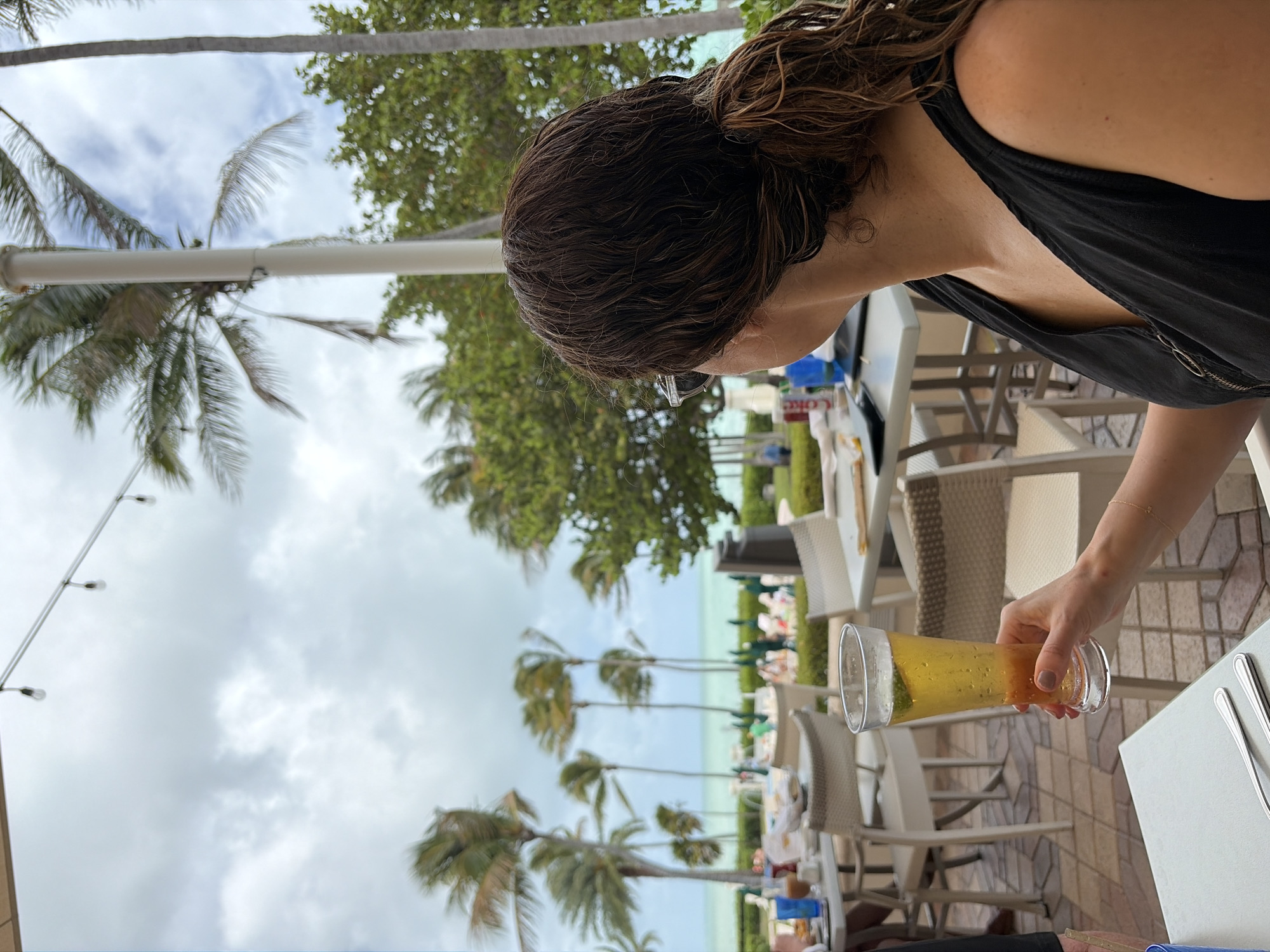 The photograph shows a woman at an outdoor dining area, likely in a resort or upscale casual restaurant, in what is determined to be the city of Key Biscayne, United States of America. The woman has dark hair, is wearing sunglasses and a black top and she is holding a glass of beer. The drink is light amber in color, with visible bubbles, and a lime wedge is visible in the glass.
The dining area features white tables and wicker chairs. In the background, palm trees are visible, along with the ocean. The presence of palm trees and the ocean indicates a tropical or subtropical setting consistent with Key Biscayne. The lighting suggests a daytime shot with slightly overcast conditions.