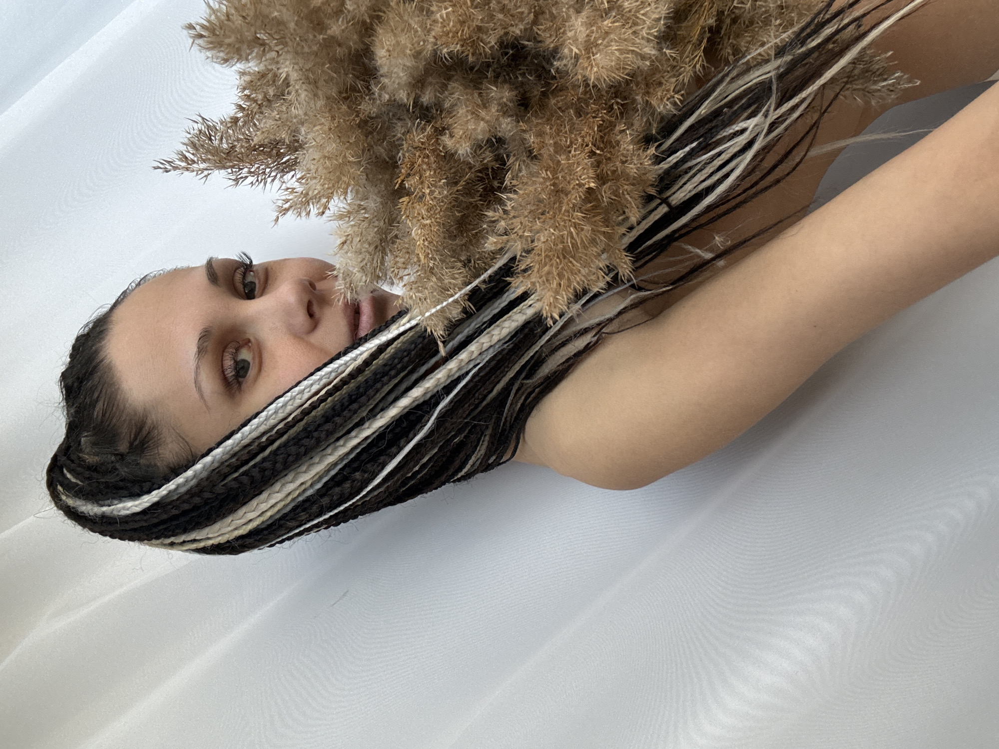 A medium shot of a woman lying on her side on a white surface. The woman has fair skin, brown eyes, and dark eyebrows. Her hair is styled in black and white braids that frame her face. She is holding a bouquet of pampas grass near her face. The background is plain white, possibly a sheet or fabric. The scene appears to be indoors. No specific information indicates the location is Rusanovo, Russia.