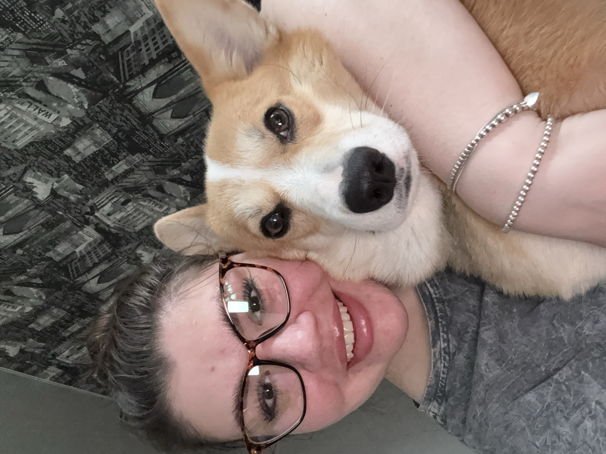 A woman and a Corgi dog are close together in an indoor setting. The woman is wearing glasses and smiling, while the dog has its head near the woman's face. The woman is wearing a bracelet on her arm. The background shows a patterned surface, possibly a couch or bedding. The location is unknown.