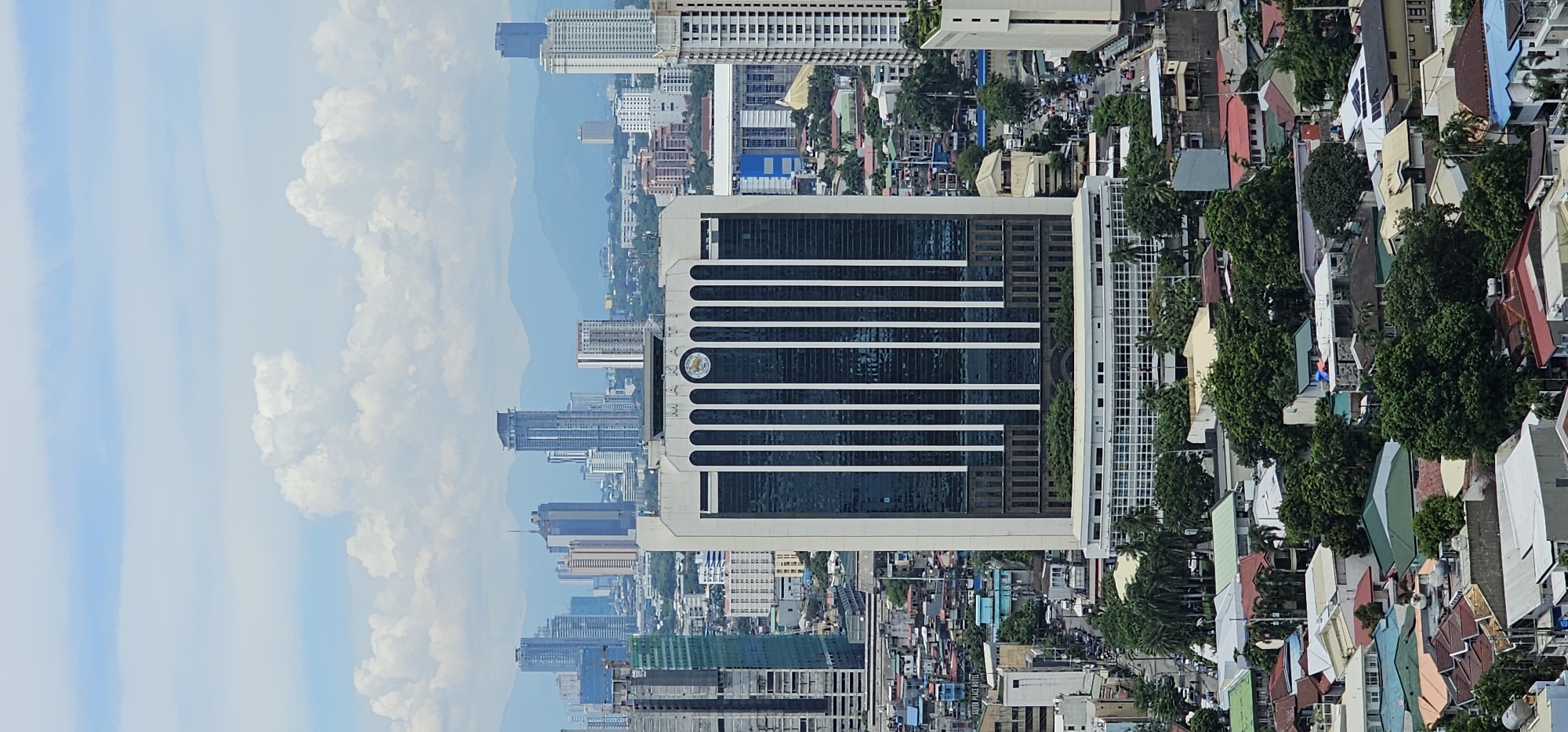 This is a vertical, high-angle, full-shot photograph of a cityscape in Makati, Philippines. The image is dominated by a large, rectangular, white and glass office building in the center, with "MAKATI" emblazoned in capital letters across its upper facade, along with a circular emblem to its right. The building features a repeating pattern of vertical white panels against dark glass. Below this main building, the image shows a lower, white, multi-story structure with numerous windows.
The foreground is filled with a dense collection of residential buildings with varied roof colors including red, green, and beige, interspersed with lush green trees. A street with some vehicular traffic is visible to the right of the central building.
In the background, a sprawling urban landscape unfolds with numerous other skyscrapers and buildings, suggesting a dense metropolitan area. Beyond the city, hazy blue mountains form the horizon under a sky filled with scattered white clouds against a light blue backdrop. The overall impression is that of a typical day in a large, developing Asian city, with a mix of modern architecture and older residential areas. There are no visible people in the frame.