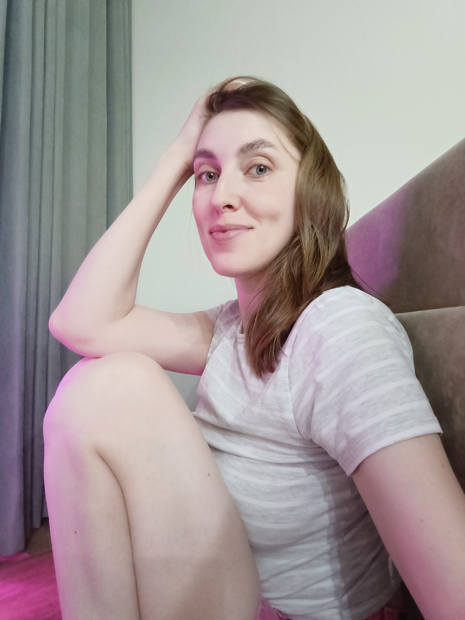 A woman is captured in a medium-close-up, relaxed portrait, likely taken indoors in Chaikovsky, Russia. She is positioned with her body turned slightly to the right, facing the viewer directly with a soft, gentle smile. Her head is tilted slightly, and her right arm is bent, with her hand resting casually on her forehead or hair. She has light brown, shoulder-length hair and light-colored eyes.
She is wearing a light-colored, possibly grey and white, horizontally striped short-sleeved t-shirt. Her legs are bent, visible in the lower part of the frame, suggesting she might be sitting on the floor or a low surface.
The setting appears to be a room, with grey, vertical curtains visible on the left side of the image. On the right, part of a brown, upholstered piece of furniture, possibly a sofa or chair, is visible. The lighting in the scene is notable; there is a distinct pink and purple glow originating from the lower left, casting a vibrant hue on the curtains, the floor, and parts of the woman's skin and clothing. This suggests artificial mood lighting, possibly an LED light source. The overall atmosphere conveyed is calm and contemplative. No text is visible in the image.