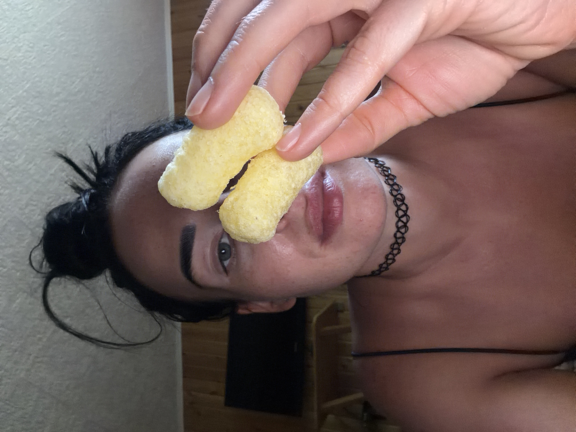 The image shows a close-up, chest-up shot of a woman holding two pieces of puffed corn snack near her face. She has her dark hair pulled up in a bun with some strands loose around her forehead. She is wearing a black choker necklace and a thin-strapped top. Her skin appears flushed. In the background, a television screen is visible mounted on a wooden paneled wall. The scene is likely indoors. There is no specific indication of the city of Sochi, Russia, within the image itself.