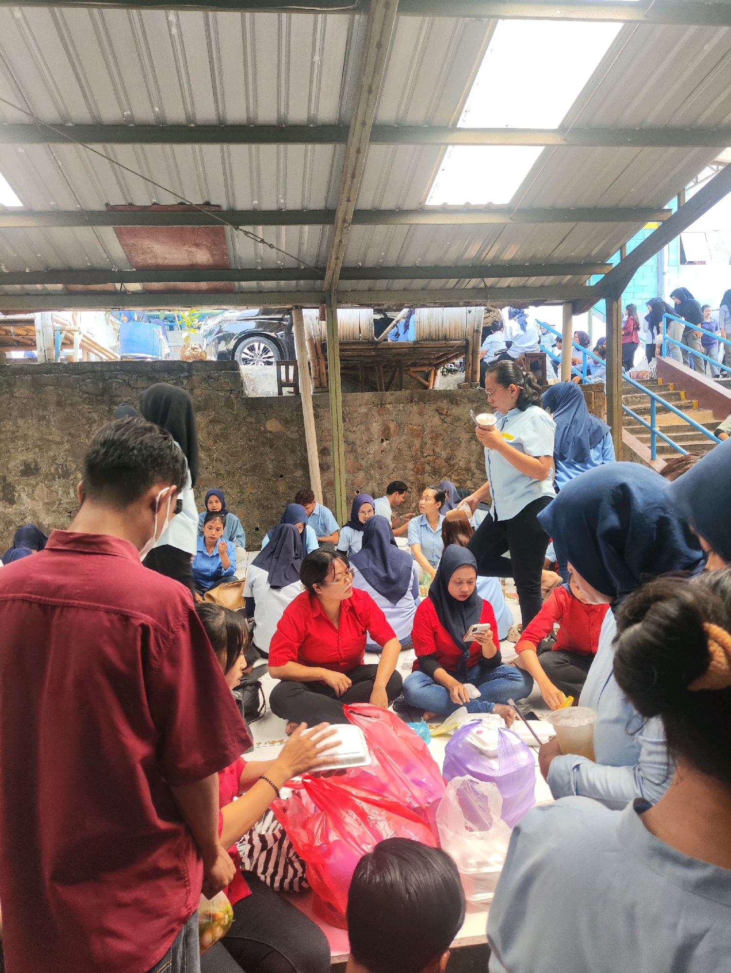 A group of people, likely students and staff, are gathered under a corrugated metal roof structure. Many are seated on the ground, and some are in casual conversation. The scene appears to be an outdoor event or a casual mealtime, possibly at a school or community gathering. Some individuals are holding food containers or drinks, and red and pink plastic bags are visible, suggesting the distribution of food or supplies. The environment includes a concrete wall behind the seated individuals and a car visible in the background. The presence of a car and the architecture suggest an urban or semi-urban setting in Randugunting, Indonesia.