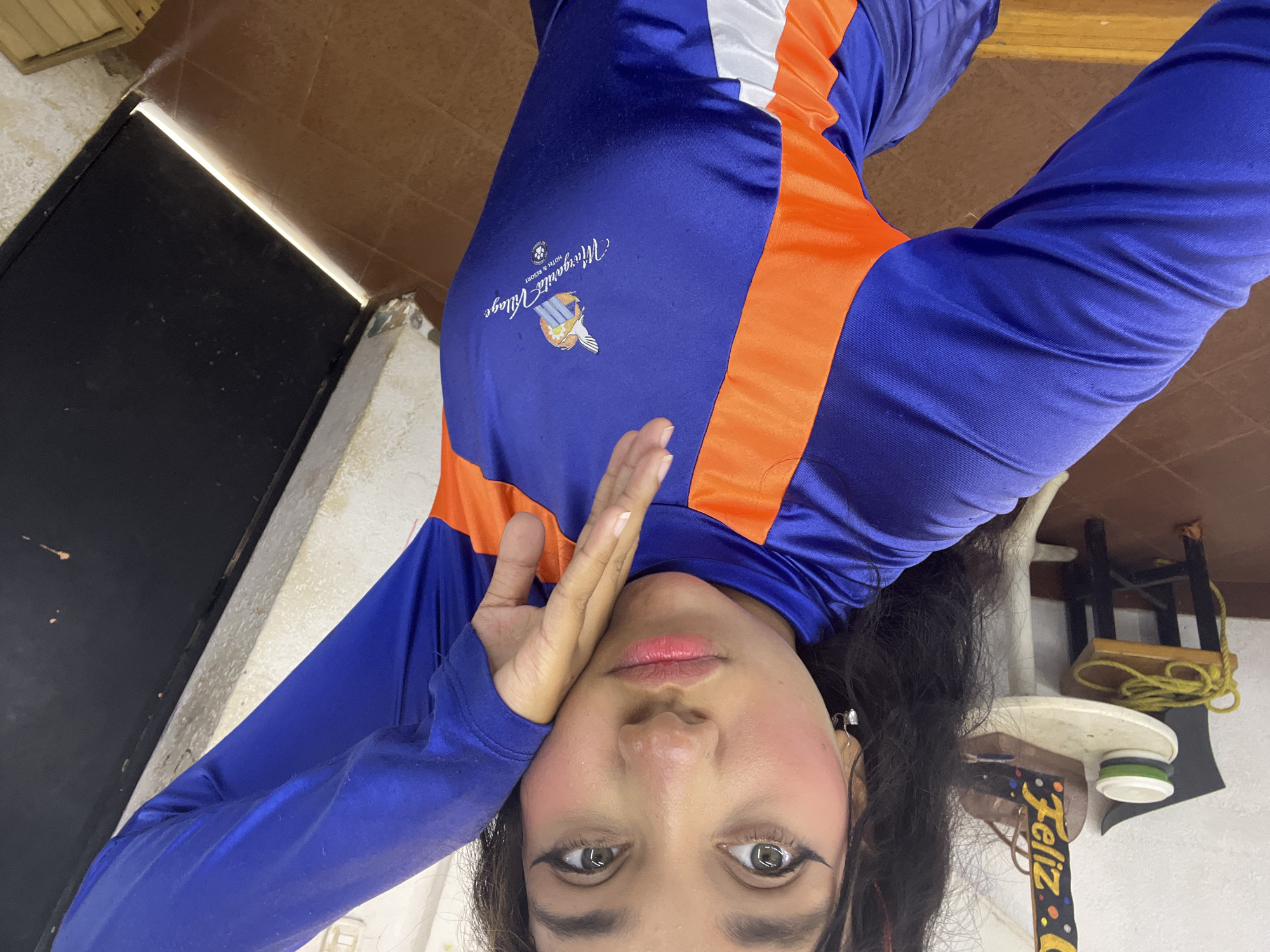 A person, seen from an upside-down perspective, is wearing a blue and orange long-sleeved shirt with the logo "Margarita Village Hotel & Resort". The individual is holding their hand up to their face. The background reveals a tiled floor, a dark door, and a table with various items on it, including what appears to be rope, stacked cups, and a decorative banner with the word "Feliz." The "Margarita Village Hotel & Resort" logo suggests that this photo was taken in Porlamar, Venezuela, as that is its location.