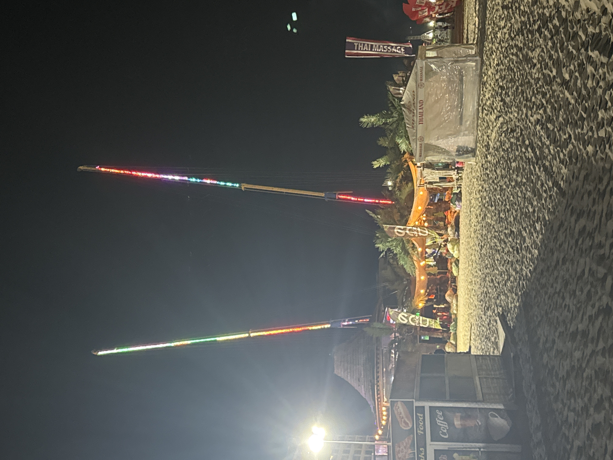 The image depicts a nighttime scene on a sandy beach, likely in Nesebar, Bulgaria, judging by the context provided. Dominating the center of the frame are two tall, illuminated amusement park rides, possibly a slingshot or bungee jump attraction, with strings of colorful lights running up their masts. To the left, a food stall is visible, displaying images of pizza and a hot dog, with "Coffee" also advertised. Behind the food stall, a large building with a thatched roof and a bright spotlight is present. Further back, tents are set up, one of which is clearly labeled "THAILAND MASSAGE". Palm trees and other vegetation dot the landscape. The sand is textured and casts shadows from the artificial lights. The sky is dark, indicating it is night. A few indistinct figures can be seen in the background, suggesting the presence of other people enjoying the area. The overall atmosphere is that of a lively beachside entertainment area at night.