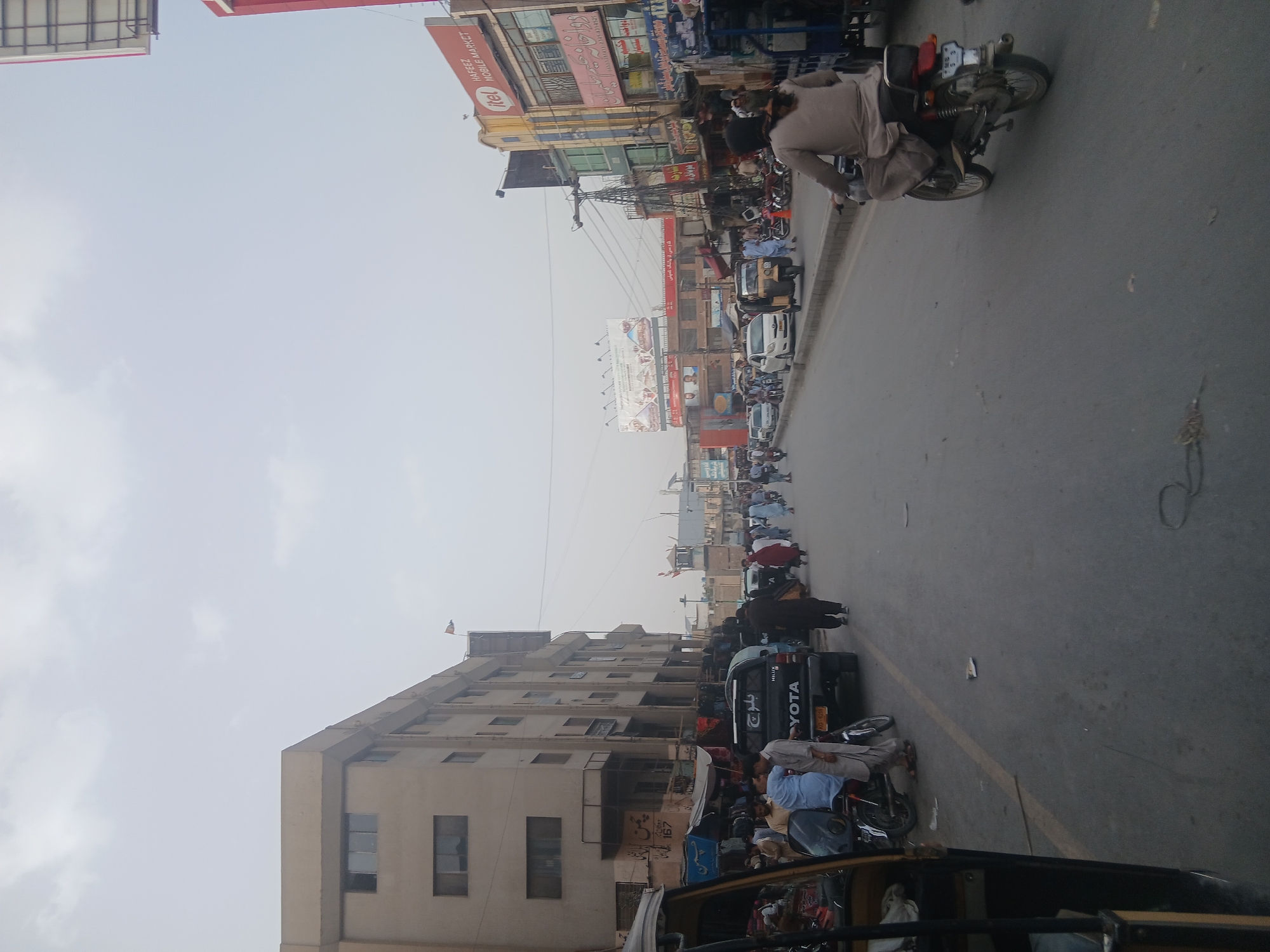 The image depicts a street scene in Quetta City Tehsil, Pakistan. A mix of pedestrians, motorcyclists, and auto-rickshaws fill the street. Buildings line both sides of the road, with shops and commercial signage visible. Notable signage includes "HAFEEZ MOBILE MARKET," "YOTA," "MILUX," and local Urdu text. The sky is overcast, and the overall scene suggests a bustling urban environment.