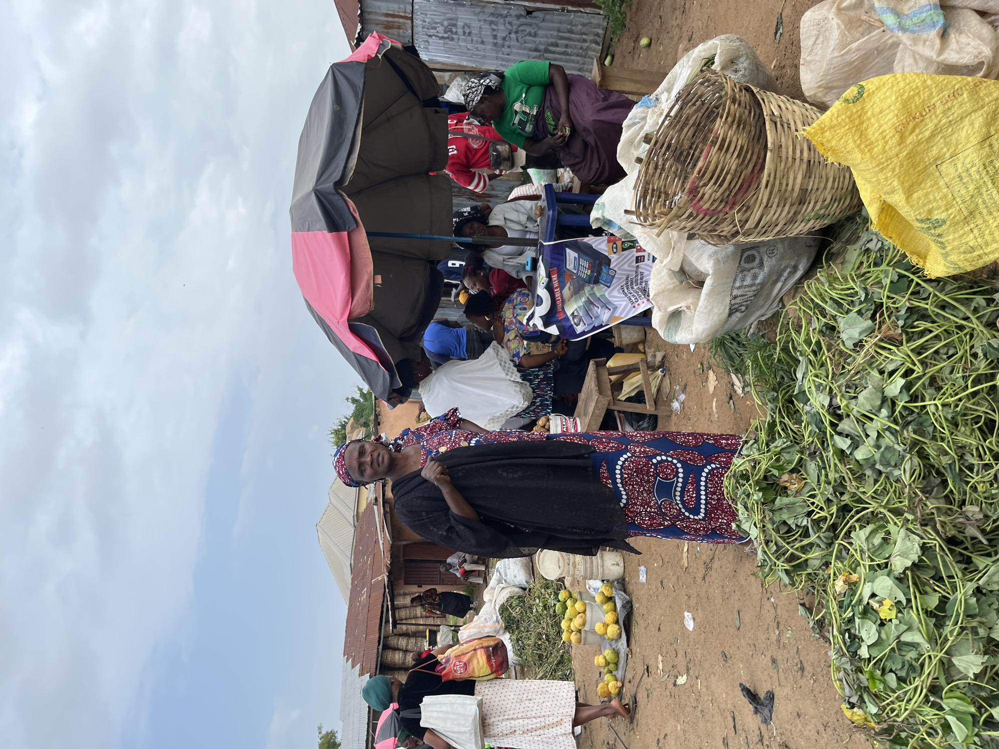 This media file depicts an outdoor market scene in Gura Top, Nigeria. The central subject is a woman standing and looking towards the camera, dressed in a black garment over a patterned blue and red dress, with a matching headscarf. She appears to be a vendor or shopper at the market.
Behind her, under a large, colorful umbrella (partially pink and dark gray), several other individuals are visible, engaged in market activities. Some are seated, seemingly interacting with goods or other people. The market appears to be set up along a dirt path, with various stalls or makeshift setups.
Objects in the scene include several large sacks, some filled with goods, one notably yellow. A woven basket, filled with what appears to be produce or other market items, is prominently placed. Piles of leafy green vegetables, likely for sale, are also visible, as are some yellow fruits. In the background, corrugated metal structures suggest the presence of shops or shelters.
The overall impression is a bustling marketplace. The sky is overcast with clouds, suggesting daytime but not necessarily bright sunshine. The ground is dusty, typical of an outdoor market. There are no explicit indications of a specific time of day beyond it being daylight. The mood is that of everyday commerce.
One visible text element is on a poster or sign partially obscured by a basket, featuring images of what looks like a point-of-sale device and currency. The text is in English and includes phrases like "WE ARE HERE" and possibly pricing or service information, suggesting a modern commercial element within the traditional market setting.