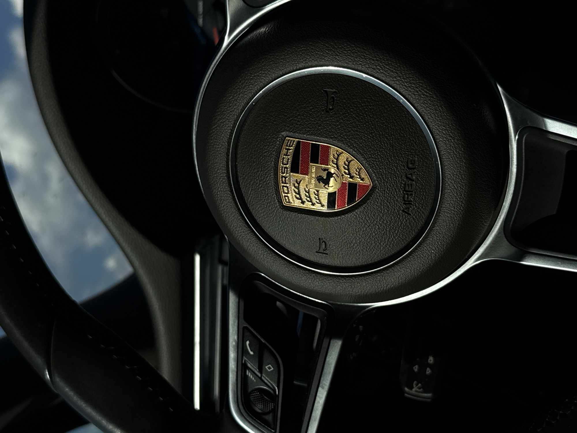 The image shows the interior of a Porsche vehicle, focusing on the steering wheel. The Porsche emblem is prominently displayed in the center of the steering wheel. Various buttons and controls are visible on the steering wheel's spokes. The surrounding interior is dark. The scene takes place in Diurtiuli, Russia.