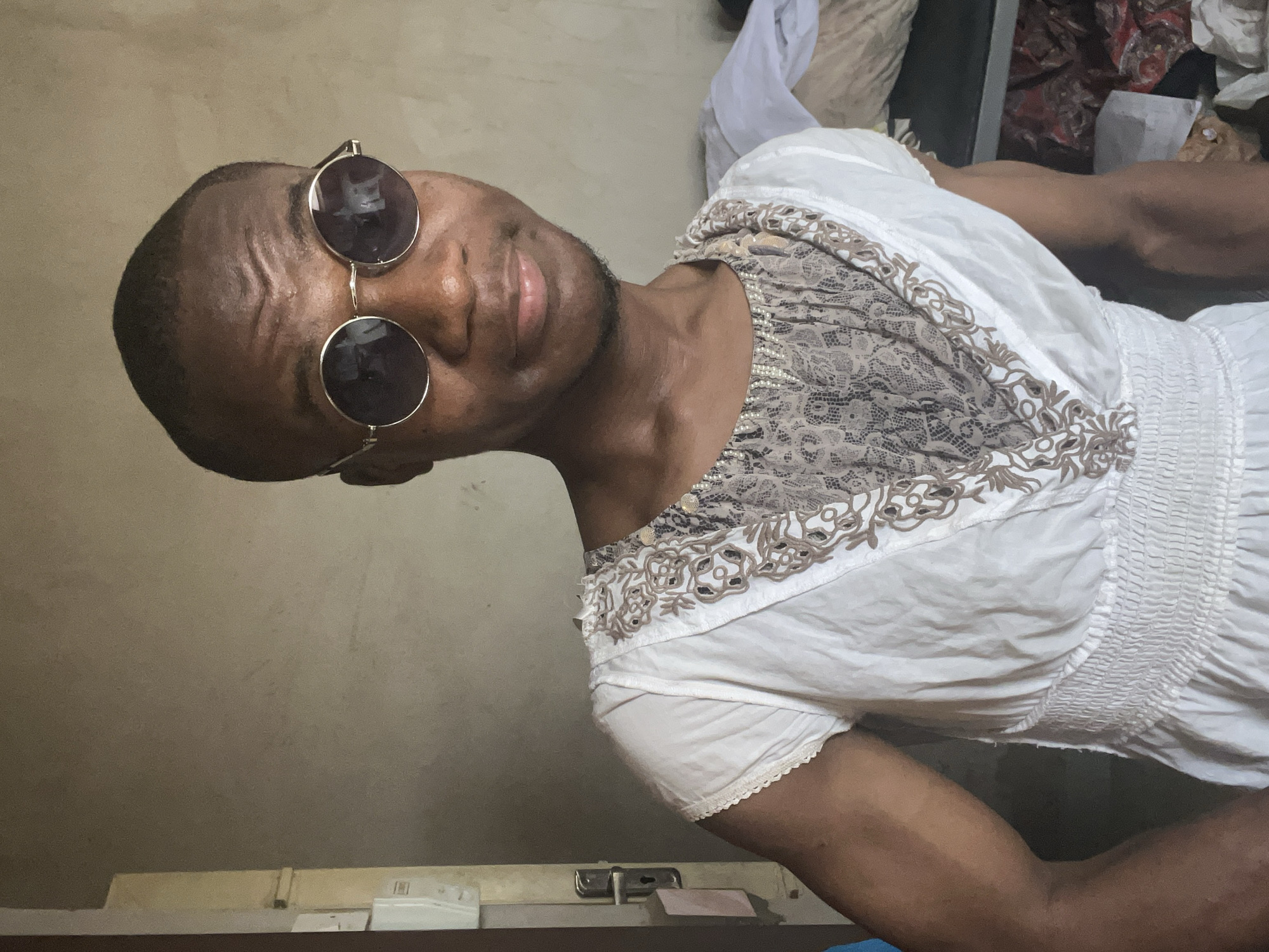 A dark-skinned person with short hair is wearing round sunglasses and a white, lacy dress. The person is posing in what appears to be an indoor setting, possibly a room in a house. The background includes a beige-colored wall. Other objects such as bedding are in the background but are not clearly visible. The location appears to be Minna, Nigeria. The photograph appears to be taken from a low angle, and the person's face is turned slightly towards the camera.