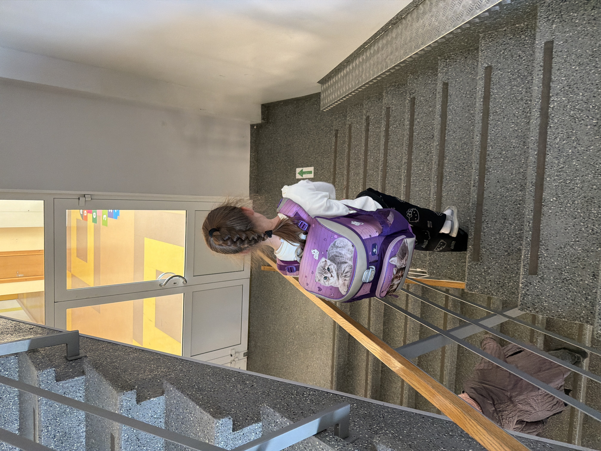 The image, oriented 90 degrees counter-clockwise, captures an indoor staircase within a building, likely a school or public institution in Riga, Latvia. The primary subject is a young girl, viewed from behind, positioned on the speckled grey concrete steps. She has light brown hair styled in a braid and is wearing a white long-sleeved top and dark pants, which display "HELLO KITTY" text. She carries a distinctive purple backpack featuring a large grey kitten on its main flap.
The staircase consists of concrete steps with a coarse, speckled finish and is equipped with metal railings, some topped with a wooden handrail. Further down the stairs, a second individual is partially visible, appearing to be seated or crouched, dressed in a brown or khaki-colored garment.
The building's interior features a combination of white walls and speckled grey walls. To the left, a large windowed section with white frames reflects vivid yellow and orange patterns, indicating brightly decorated spaces beyond. A green rectangular sign with a white arrow (pointing right if the image is correctly oriented) is visible on the grey wall, along with vertical dark grey strips and a metal grate-like structure. The ceiling is white. The scene depicts a common daily activity of traversing stairs within the building.