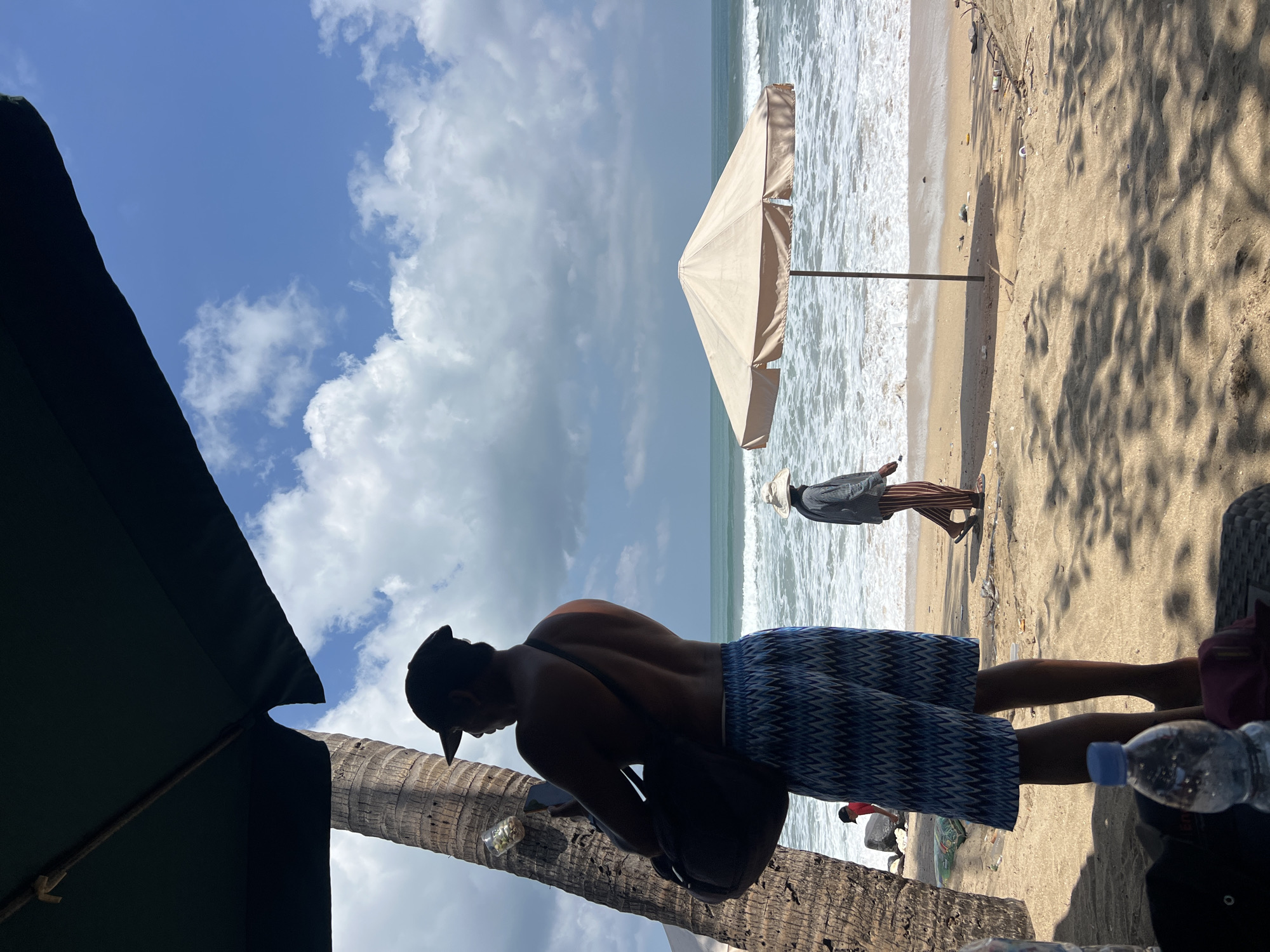 The image depicts a beach scene in Kuta, Indonesia, on a bright, cloudy day. In the foreground, a man wearing a black baseball cap and patterned blue shorts stands with his back to the camera, holding a bag over his shoulder and looking at his phone. He is partially shaded by a large dark green umbrella. To his left is a palm tree. Further back, near the water's edge, a person wearing a wide-brimmed hat and striped pants walks away from the camera, heading towards a light-colored beach umbrella. The ocean waves are visible, and the beach is sandy, with some scattered debris. In the far background, the sky is a vibrant blue with scattered white clouds.