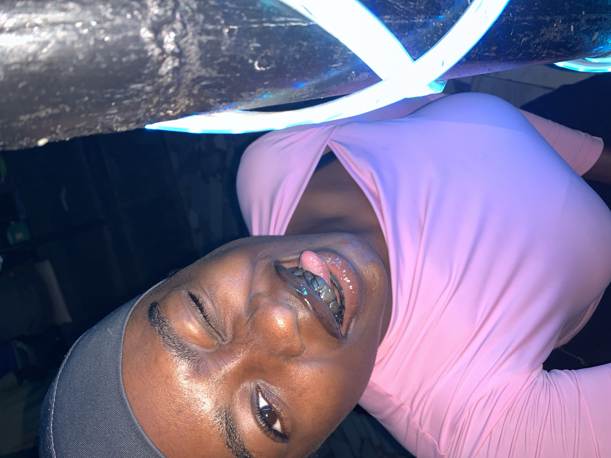 An upward-angled close-up shot depicts a person wearing a pink top with a dark head covering. The person's face is tilted, with one eye open and the other closed. Their tongue is visible, and they appear to have dental grills. Overhead, there are cylindrical structures of dark material. There is blue, possibly neon lighting wrapped around the upper objects. The location and time of the image are unknown.