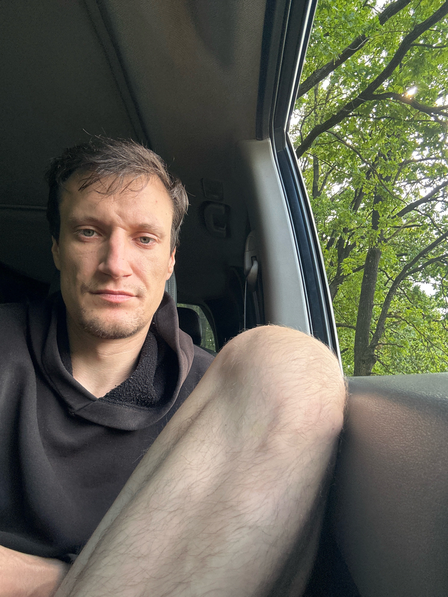 A man with light brown hair and stubble is in the driver's seat of a vehicle, looking directly at the camera. He is wearing a black hoodie. His left leg is prominently featured in the foreground, resting on the center console. The vehicle's interior is dark, with the window open to a bright, leafy green tree canopy. The image was taken in Makiv, Ukraine.
